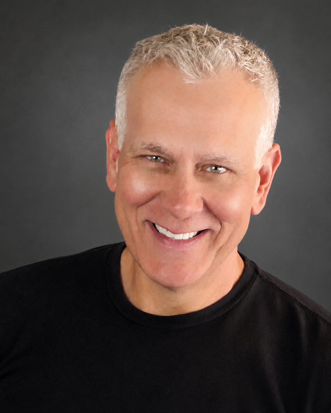 Headshot of a smiling man with short, light-colored hair and a black shirt, against a dark background.