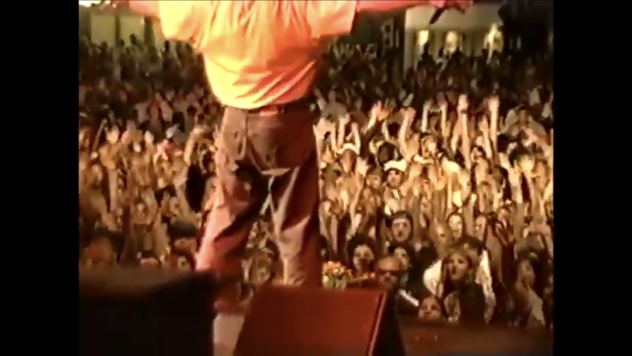 A person in a red shirt and beige pants standing in front of a large crowd of people at night.