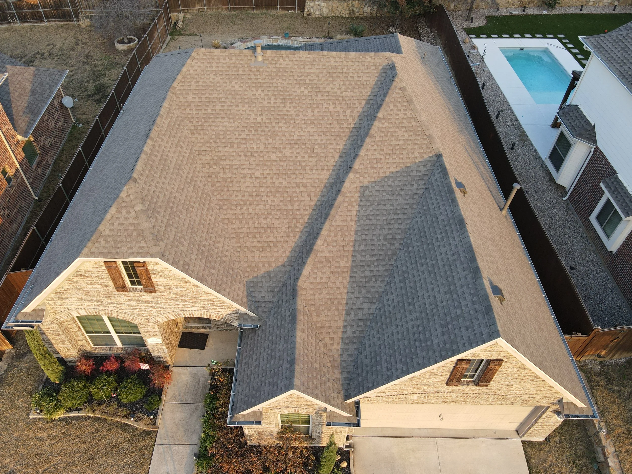 Aerial view of a house with tan and gray shingle roof, brick facade, front yard with walkway, and neighboring properties including a swimming pool in the backyard.