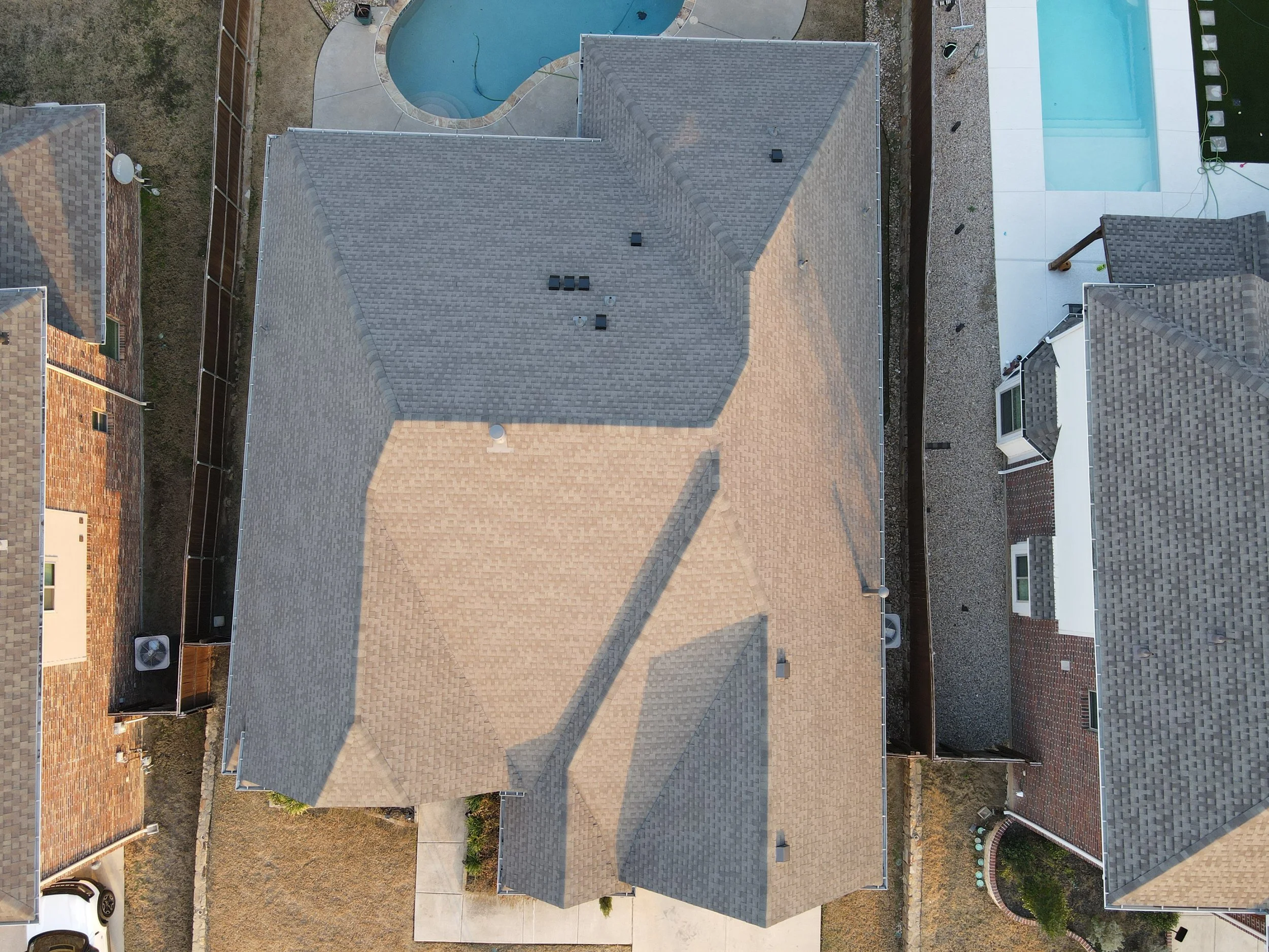 An aerial view of the roofs of multiple residential houses, one with a light gray shingle roof, a swimming pool, a white poolside patio, and a backyard with a gravel surface.
