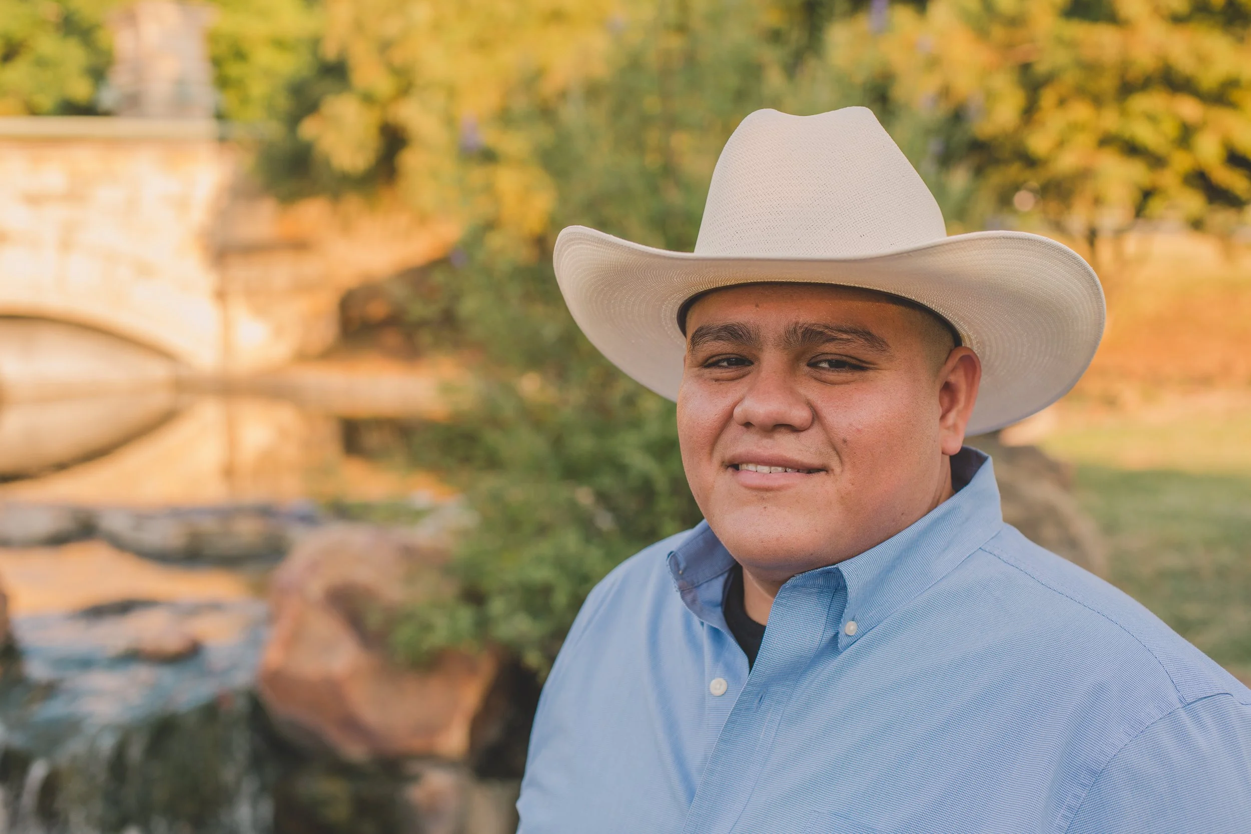 Man wearing a white cowboy hat and a blue shirt standing outdoors with trees, a bridge, and a river in the background.