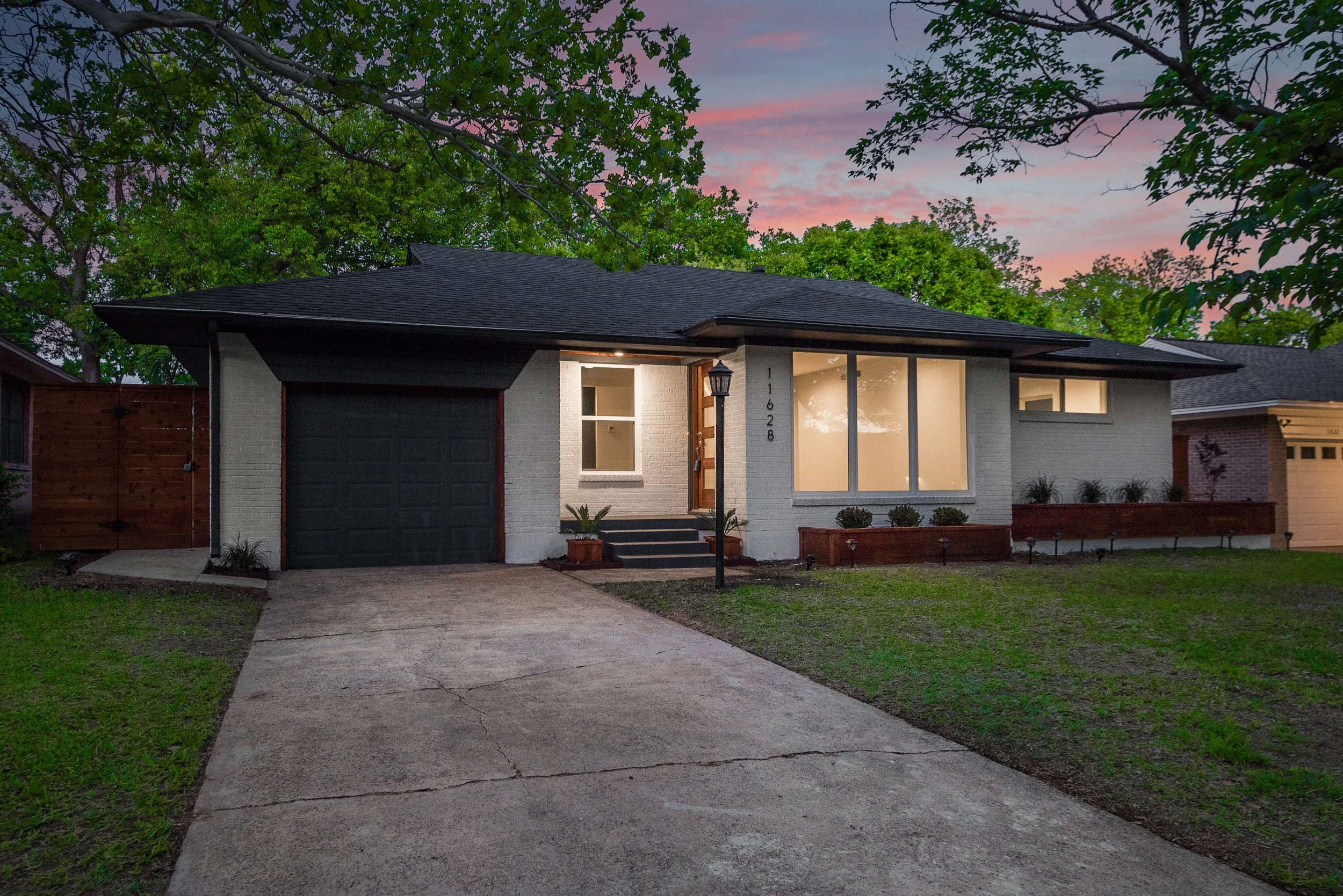 A single-story modern house with white brick exterior, black garage door, and large front windows, illuminated by exterior lights at dusk, with trees and a colorful sky in the background.
