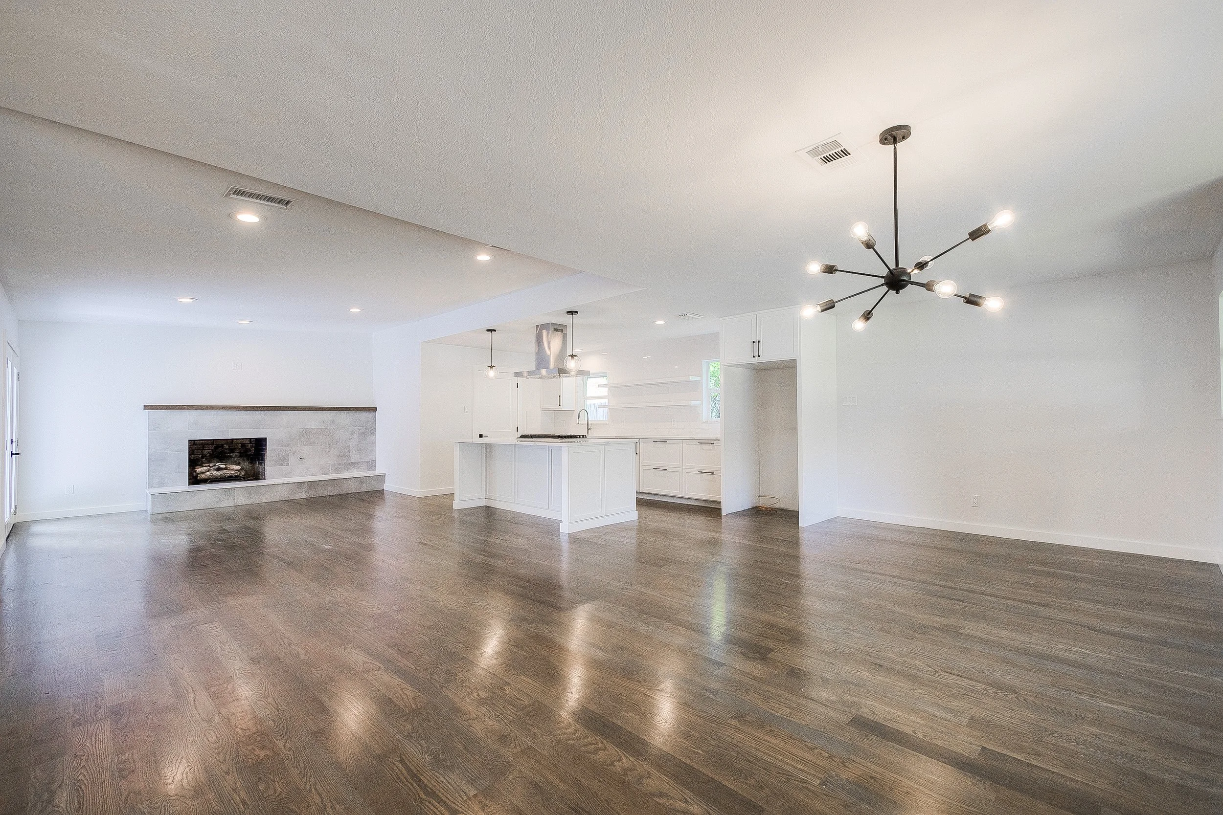 Spacious open-concept living area with hardwood floors, white walls, a modern chandelier, a fireplace, and a white kitchen with an island.