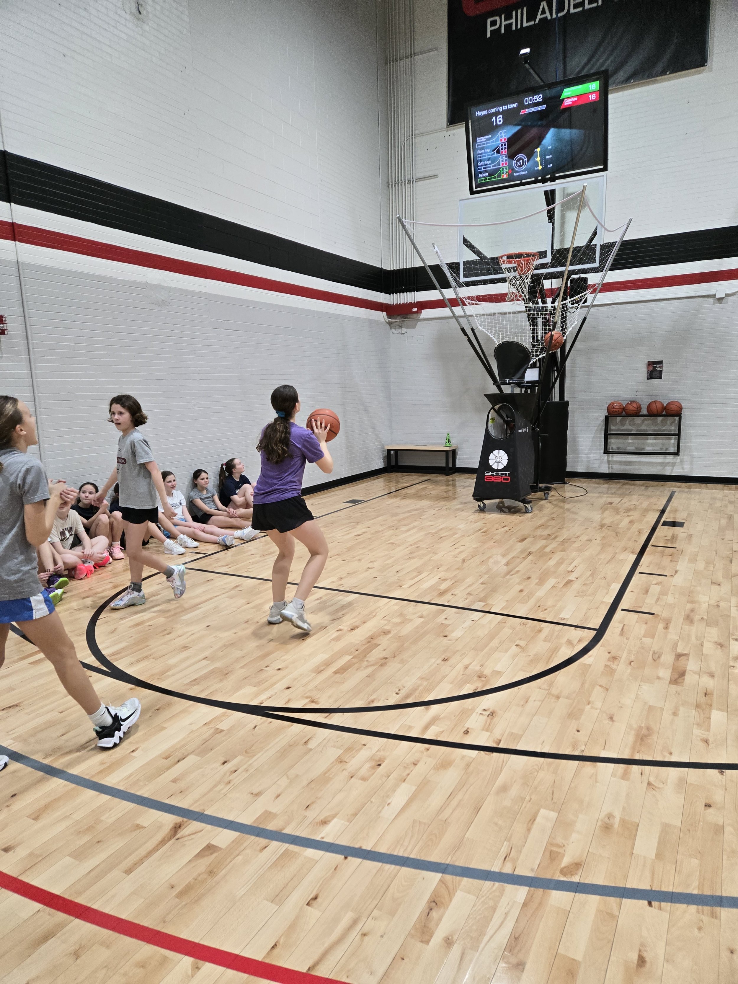 Young girls playing basketball in an indoor gymnasium, with some girls sitting on the floor watching and others actively playing near the basketball hoop.