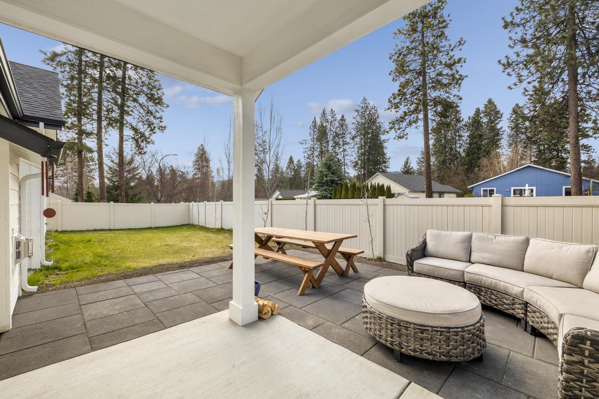 Outdoor patio with a picnic table, cushioned sectional sofa, and round ottoman on a tiled surface, surrounded by a white fence and trees.