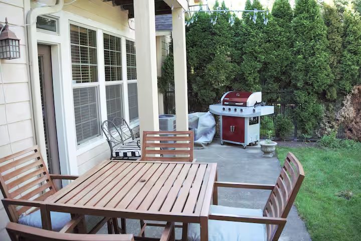 Backyard patio with wooden dining table, chairs, and a gas grill; surrounded by a hedge.