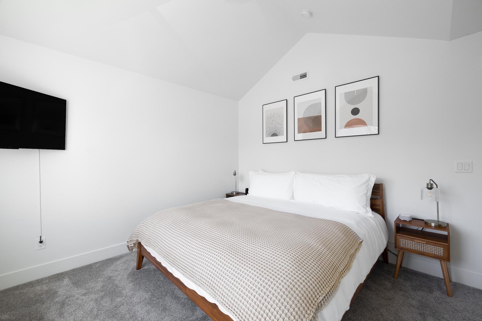 Minimalist bedroom with a king-sized bed, white bedding, taupe blanket, three framed artworks on the wall, a wooden nightstand with a lamp, and a wall-mounted flat-screen TV.
