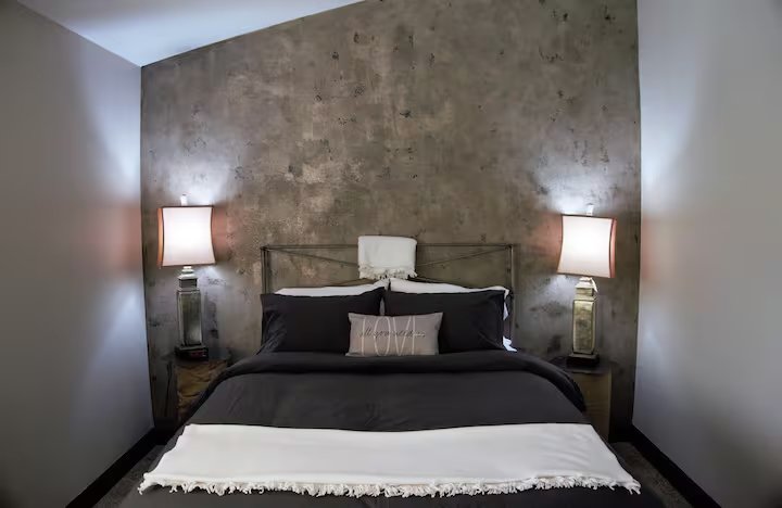 Modern bedroom with an industrial-style concrete accent wall, a cozy bed with black and white bedding, and two side tables with matching lamps.