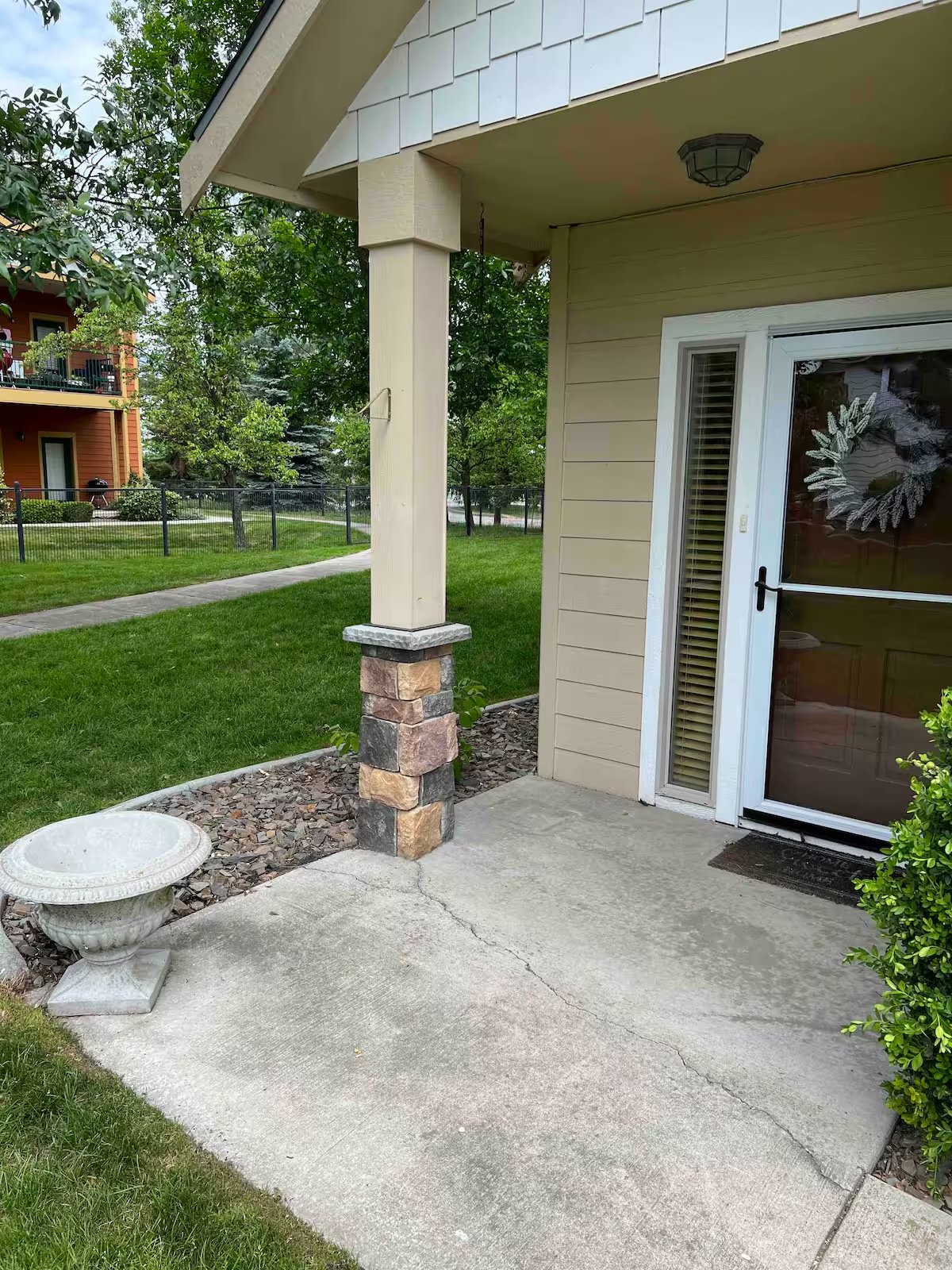 Residential porch with stone pillar, potted plant, and glass door, surrounded by greenery and a walkway.