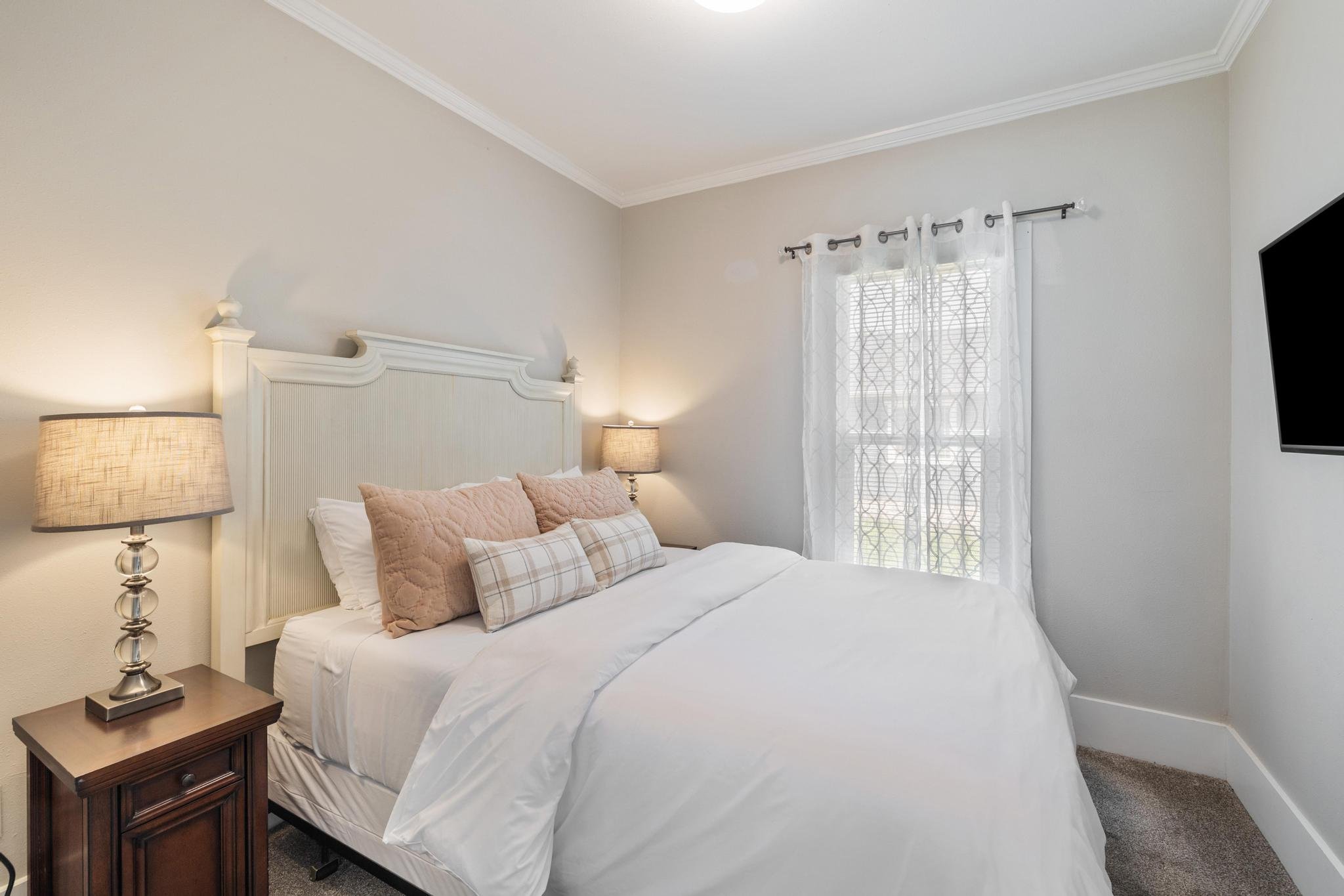 Small bedroom with beige walls, white trimmed ceiling, and a window with sheer curtains. The room features a bed with a large headboard, white bedding, and decorative pillows. Two side tables with lamps are on either side of the bed, and a wall-mount