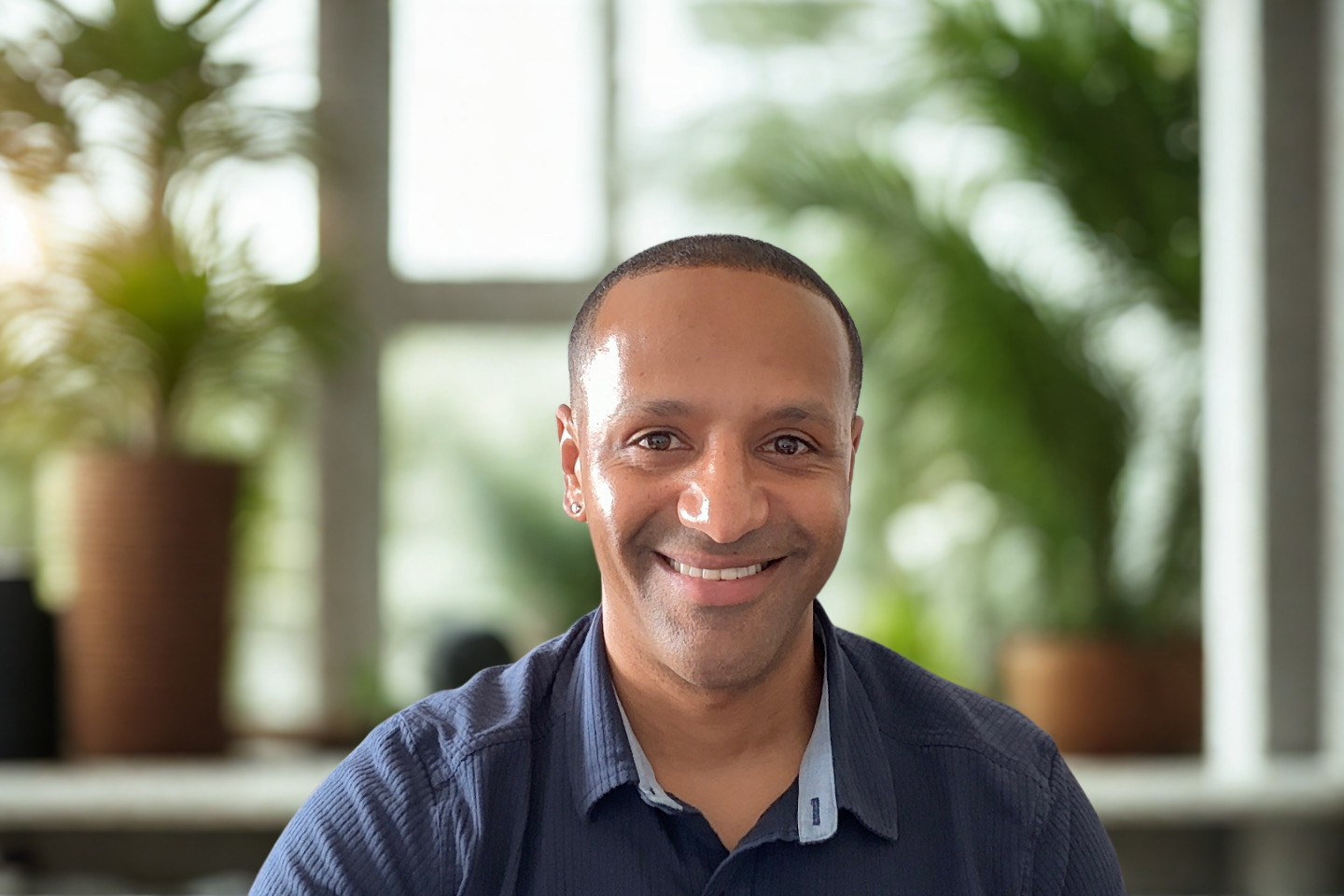 A smiling man with short hair and earrings, wearing a blue shirt, sitting indoors with blurred potted plants and windows in the background.