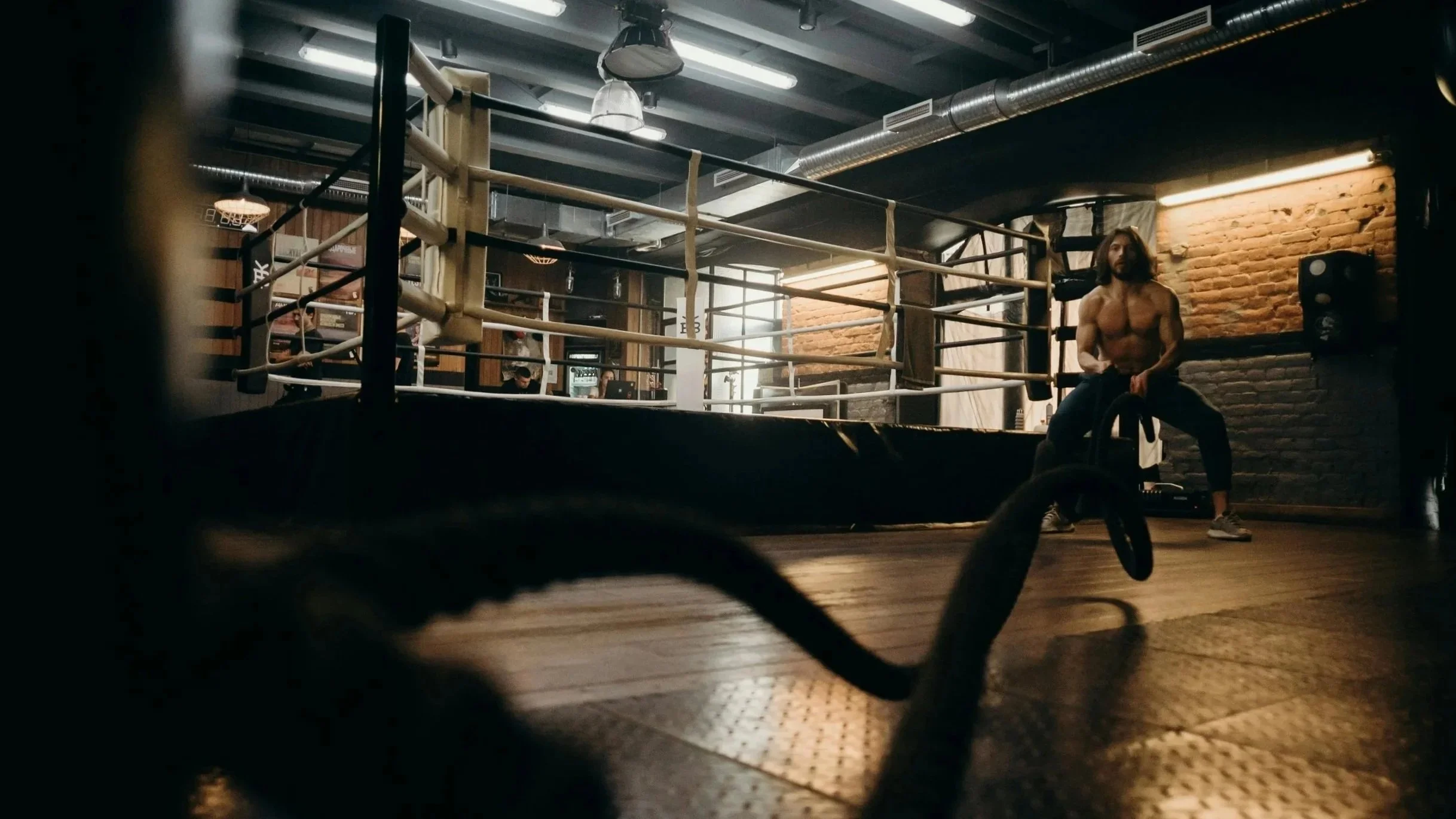 A shirtless man sitting on a boxing ring rope in a gym with brick walls and industrial lighting.