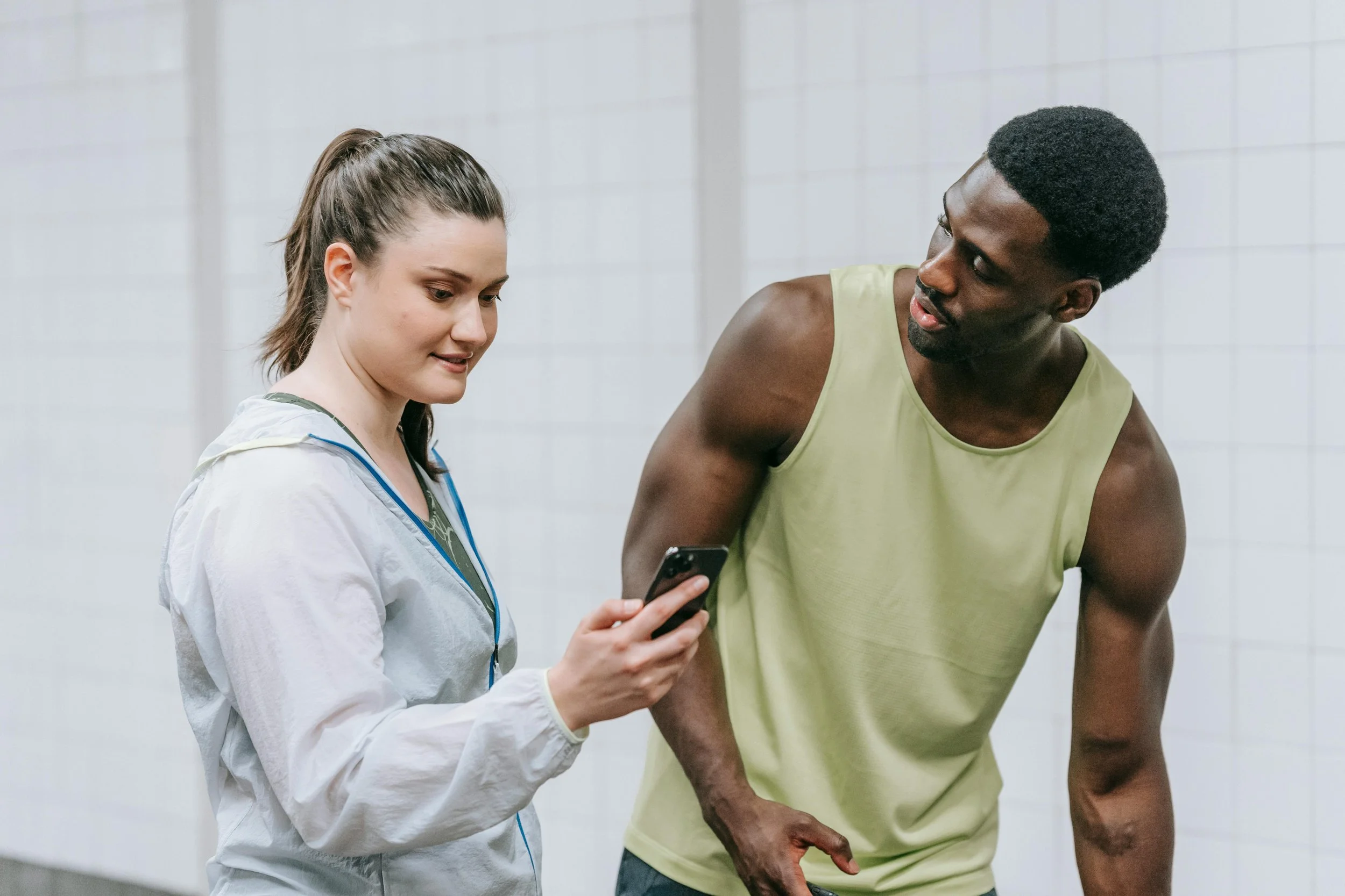 A young woman in white athletic wear shows a phone to a young man in a yellow sleeveless shirt in a gym setting with white tiled walls.