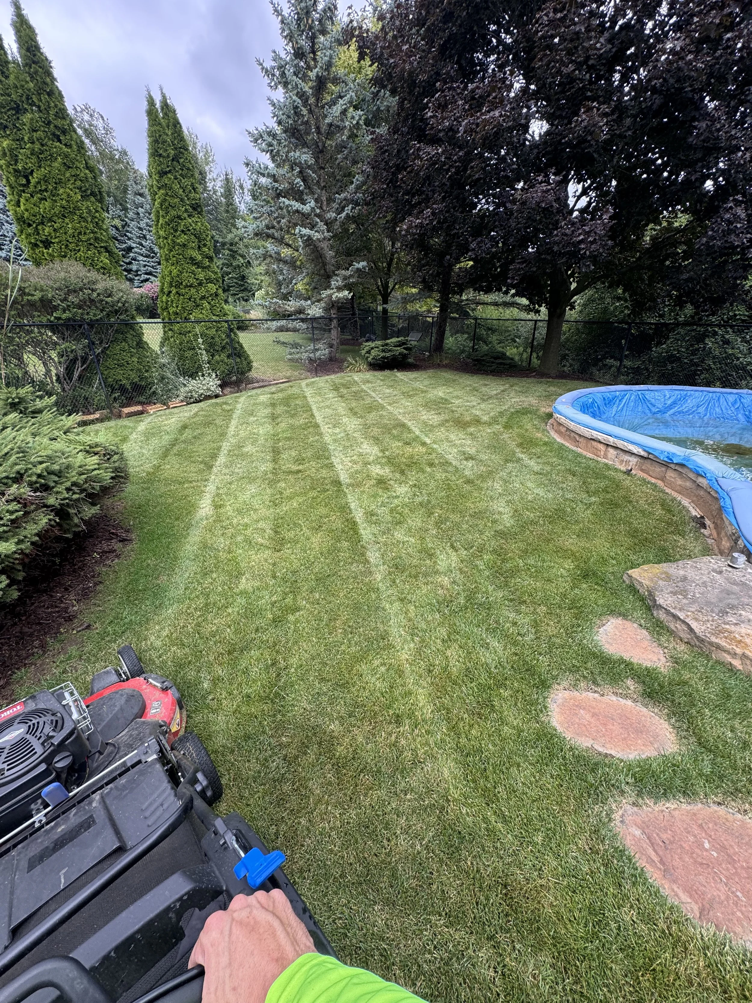 A backyard with a well-maintained lawn, garden, trees, a small inflatable pool, some stepping stones, and a lawn mower. The shot is taken from a person wearing a green shirt who is holding a lawn mower.
