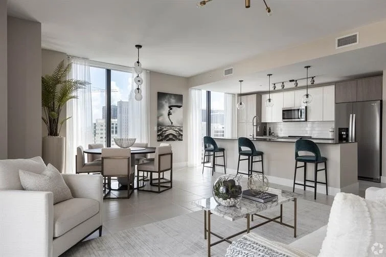Open-concept living room and kitchen with white and gray decor, a dining table with six chairs, a sectional sofa, a coffee table, bar stools at the kitchen counter, large windows with sheer curtains, and city views.
