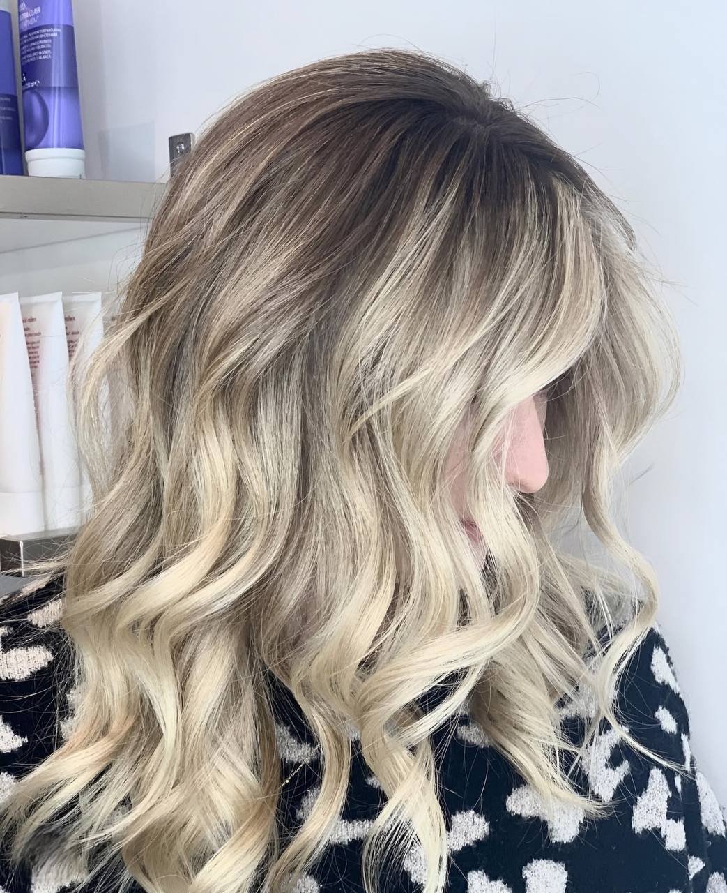 Woman with wavy blonde hair and dark roots, wearing a patterned shirt in a salon setting.