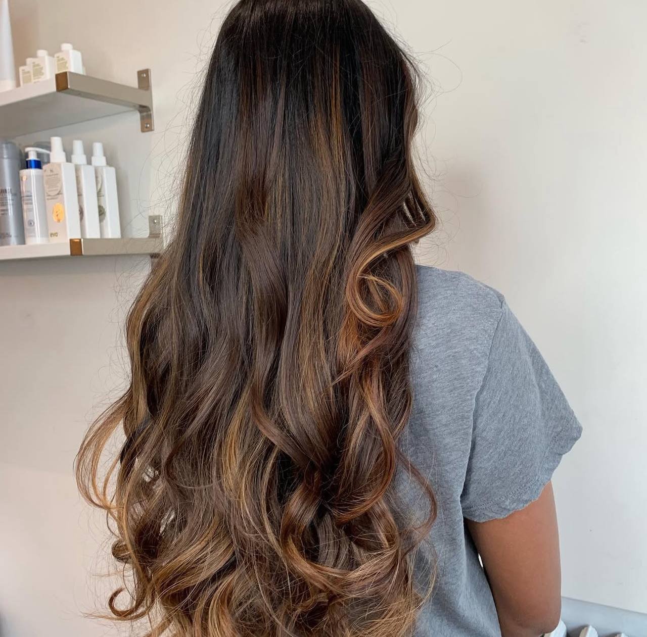 A person with long, wavy, brown hair with caramel highlights, wearing a gray top, facing away in a salon setting.