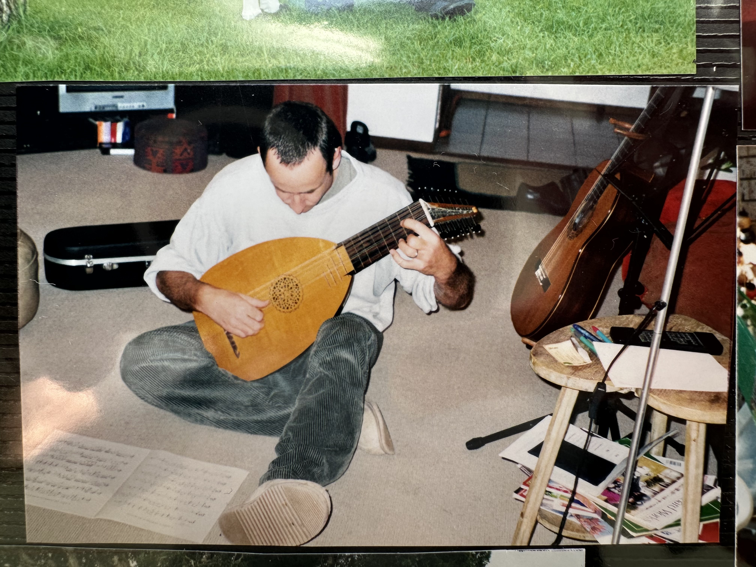 A man is sitting on the floor playing a classical guitar. There are musical sheets on the ground in front of him, and a guitar case behind him. The room has green carpet and various items, including a stool, a second guitar, and some papers.