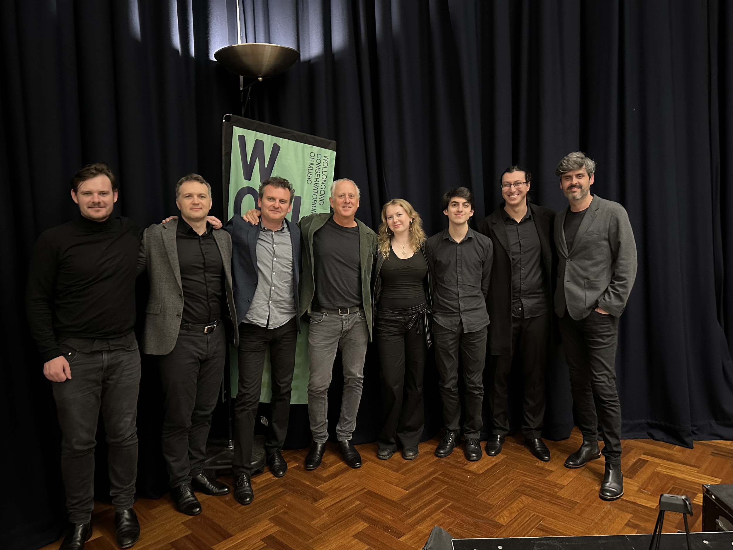 Group of seven people standing on a wooden floor in front of black curtains, smiling and posing for the photo. A green vertical banner with black and purple text is partially visible behind them, mentioning Wolfe/Wong Consulting of Music.