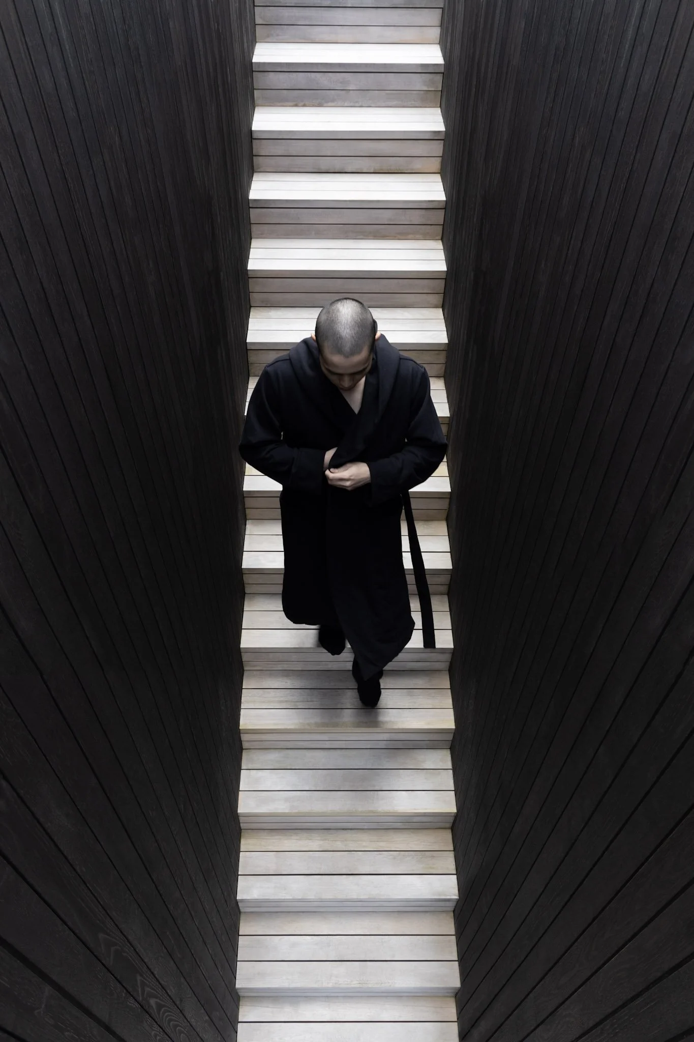 A person with a shaved head in a black robe walking down wooden stairs in a narrow, dark corridor.