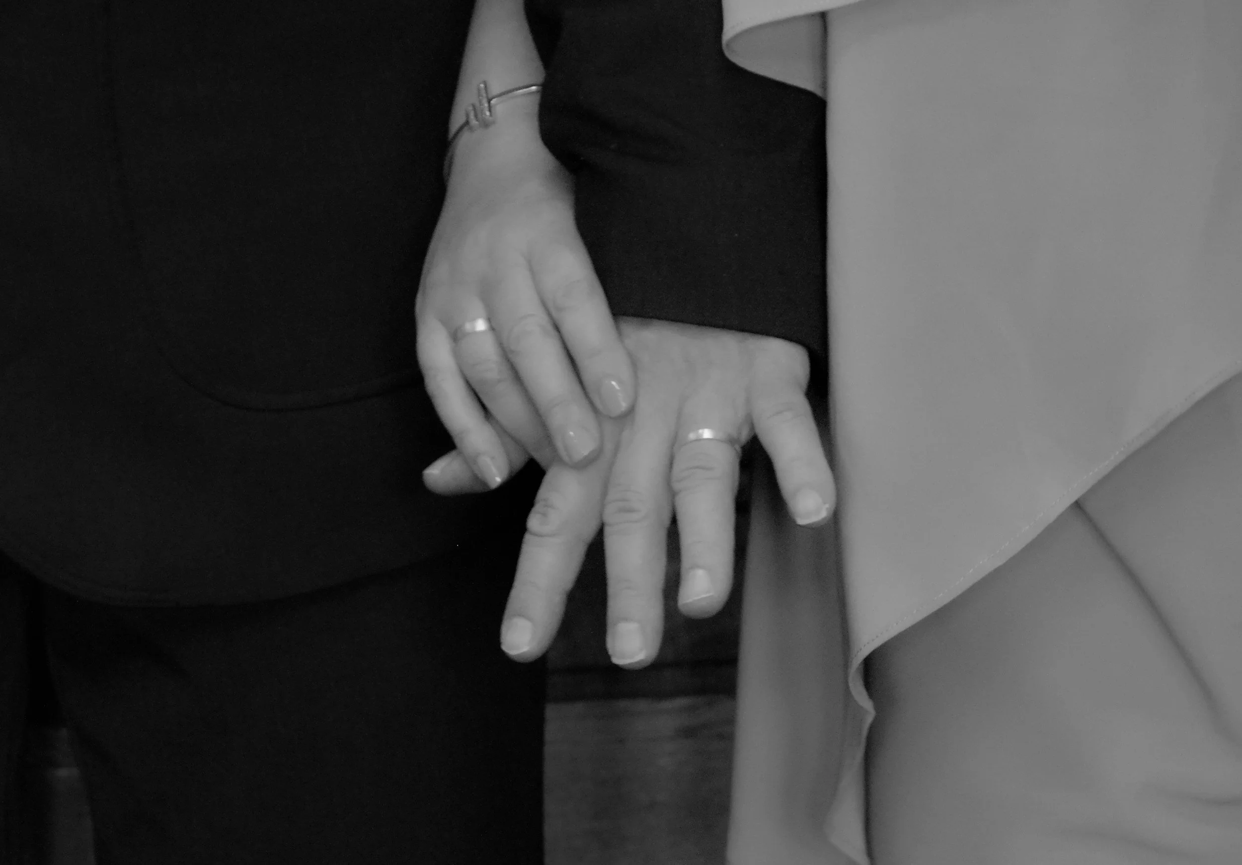 Two people holding hands, showing rings on their fingers, with a woman wearing a bracelet. The image includes parts of their bodies and clothing.