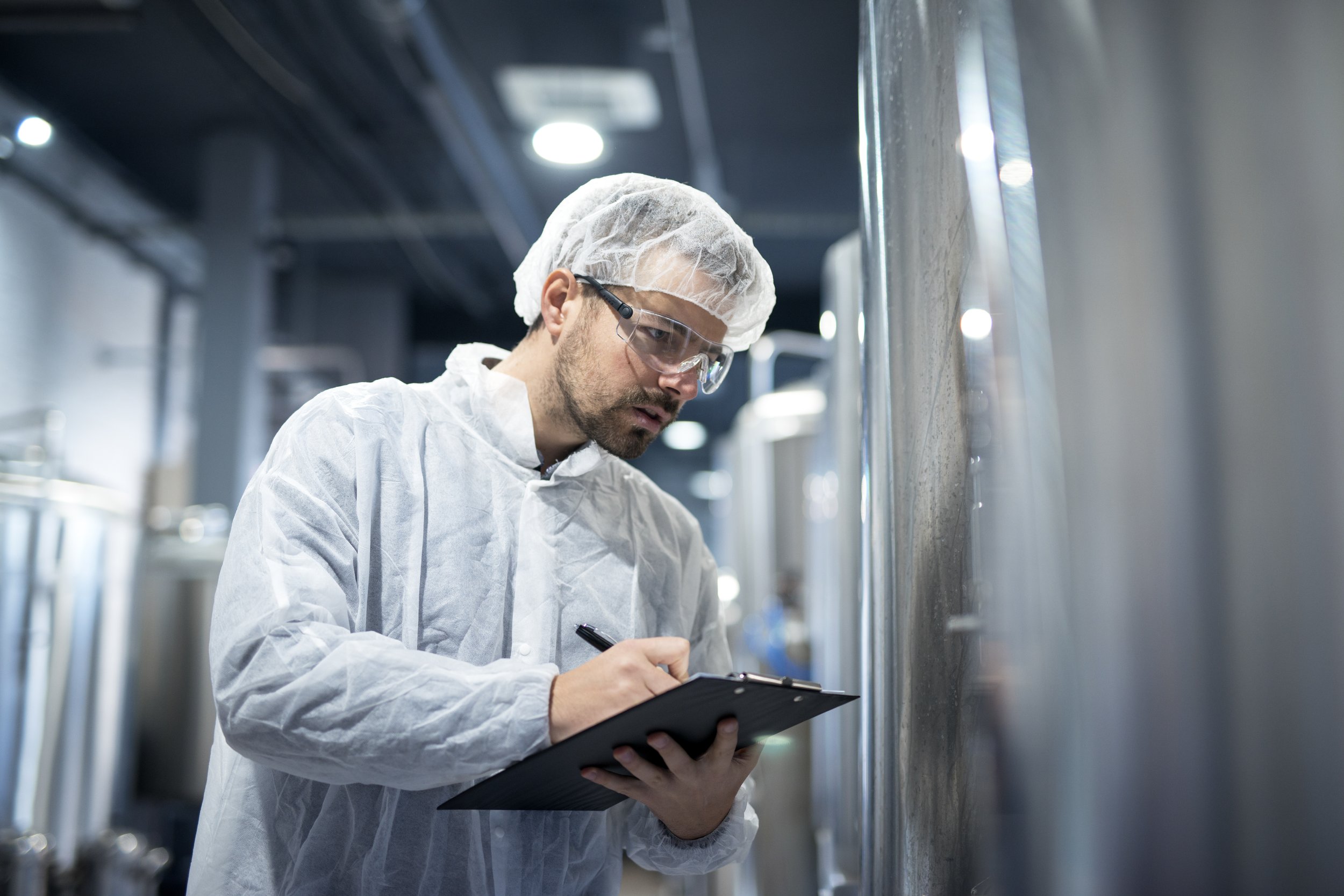 A person in protective clothing with blue gloves and a hairnet working in a clean, white industrial room filled with trays of packaged products on metal racks.