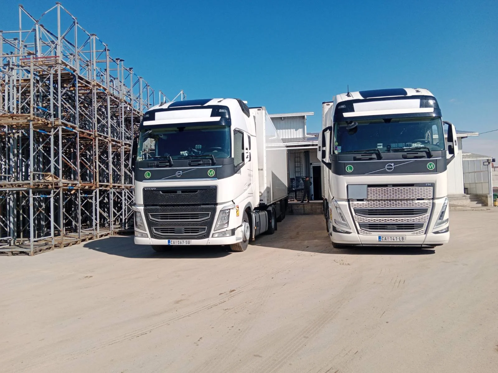 Two white semi-trucks parked next to metal scaffolding under a clear blue sky.