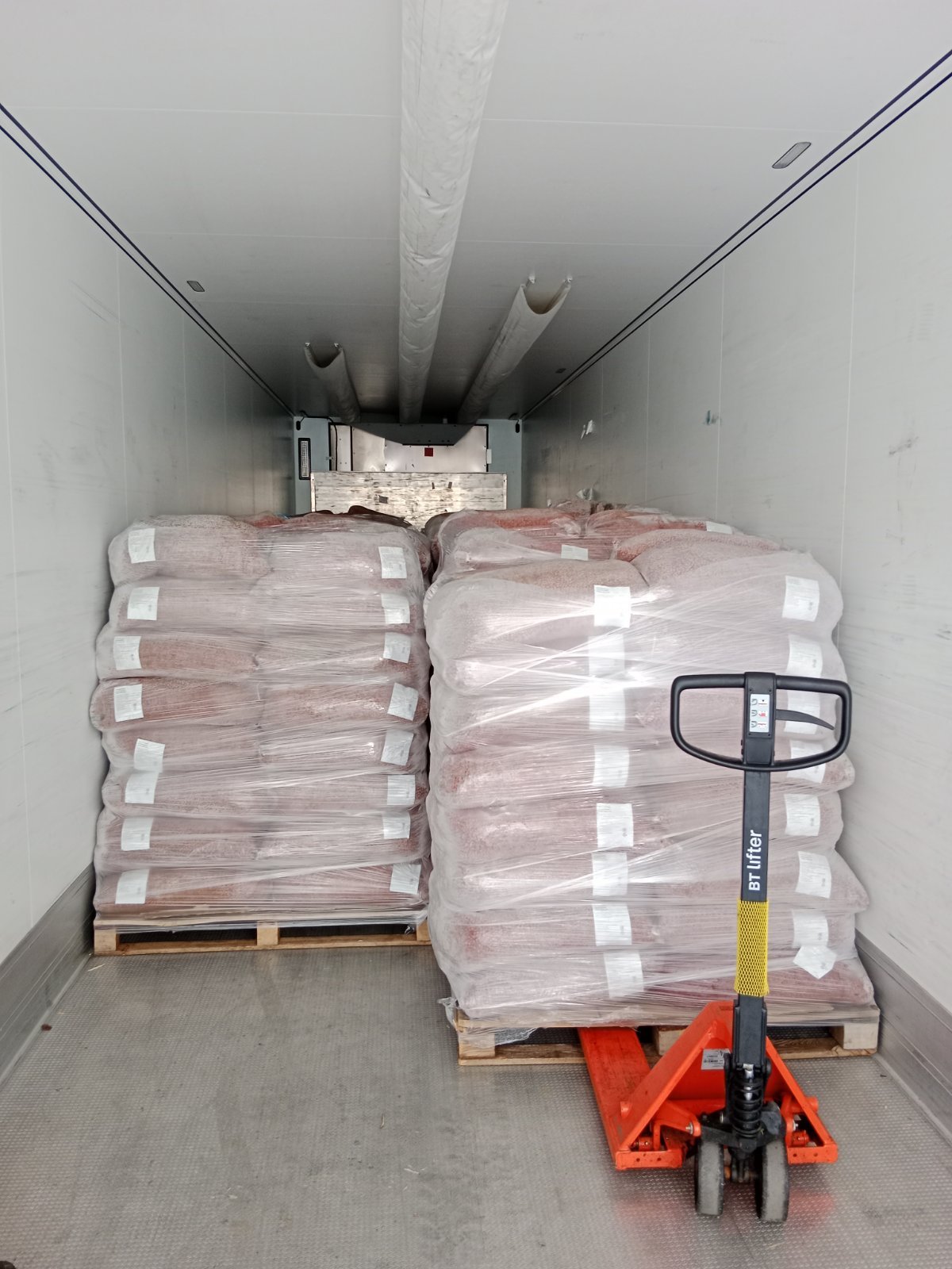 Two stacks of bagged goods on pallets in a truck with a pallet jack in front.