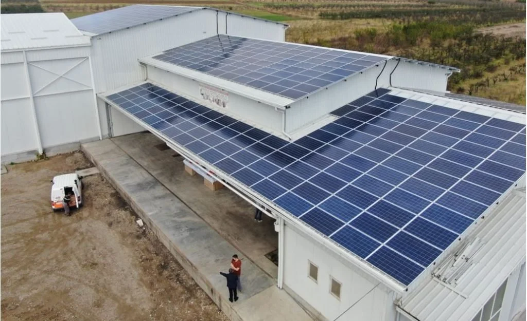 Large commercial building with multiple solar panels on the roof, located in a rural area with open land and some vegetation in the background.