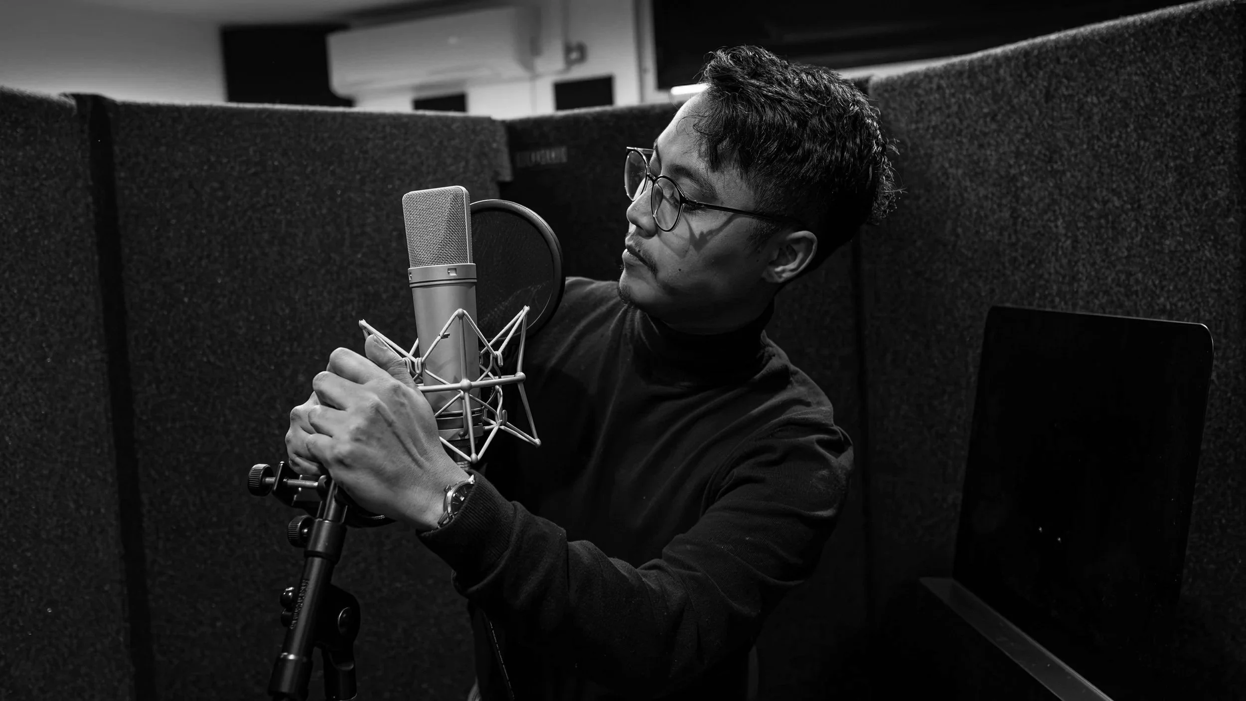 A man with glasses and a black long-sleeve shirt adjusts a microphone in a recording booth.