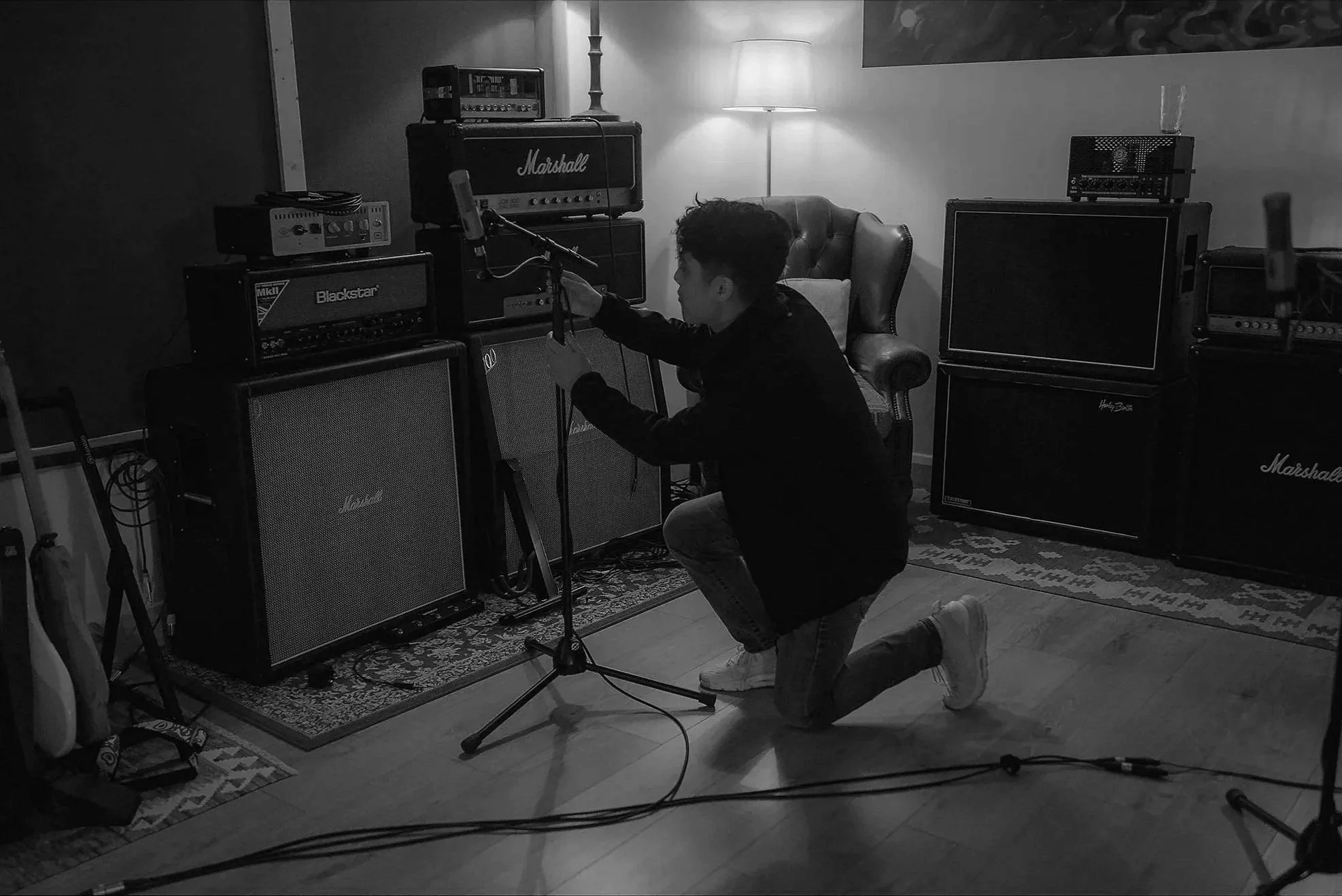 A person kneeling on one knee holding a microphone in a music studio, surrounded by guitar and amplifier equipment.