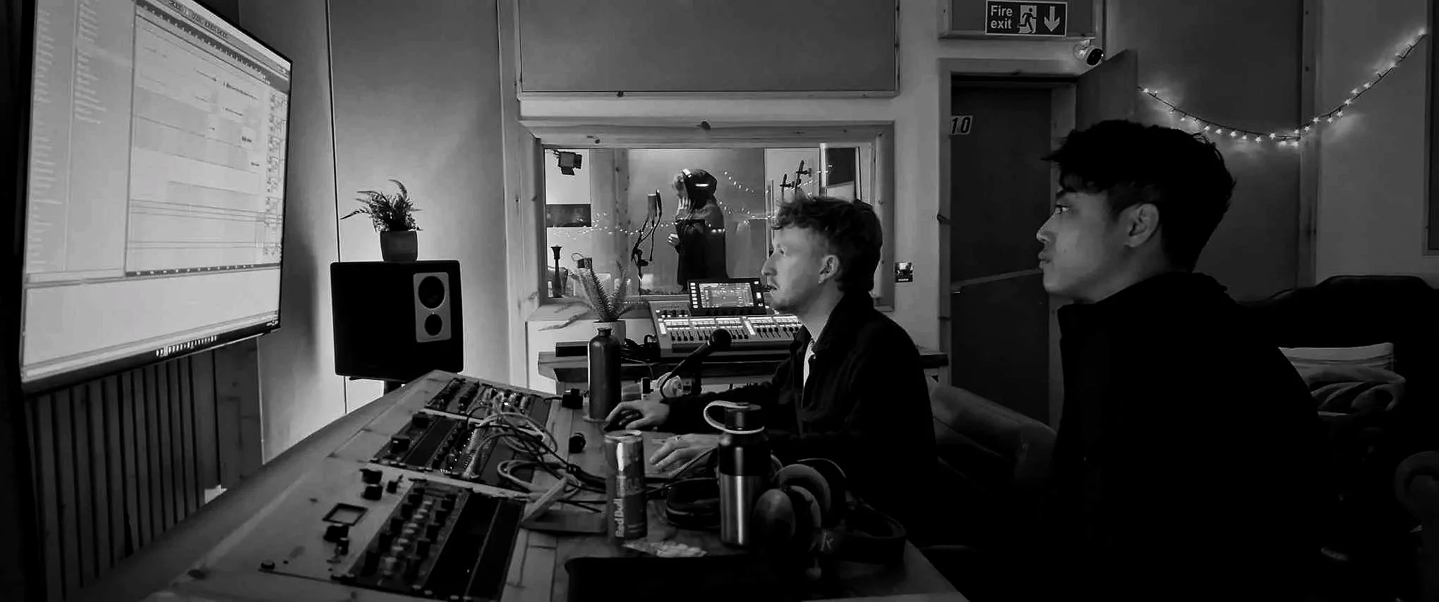 Two young men in a recording studio working with music equipment, one operating a mouse and keyboard, the other looking at the large computer screen, with a glass window revealing a woman in headphones in a vocal booth.