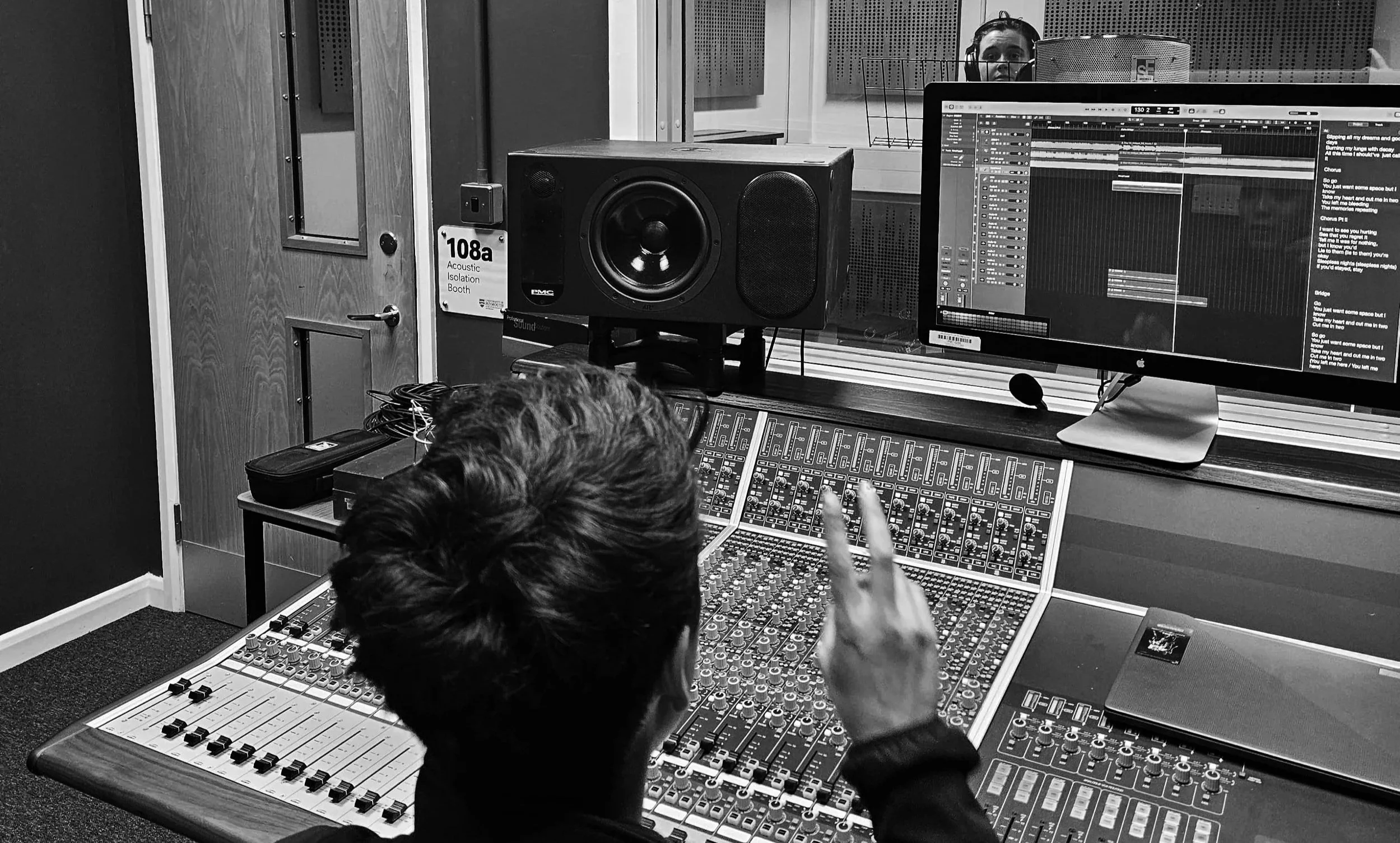 A person working in a recording studio at a mixing console, with a large monitor displaying digital audio workstations and a black speaker on the table.