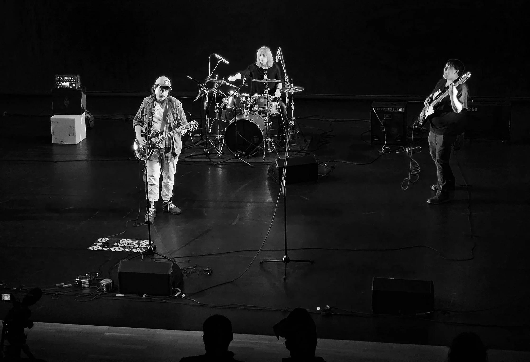 Three musicians performing on a stage: a guitarist and vocalist in the front, a drummer behind, and a bassist to the right. The scene is in black and white.