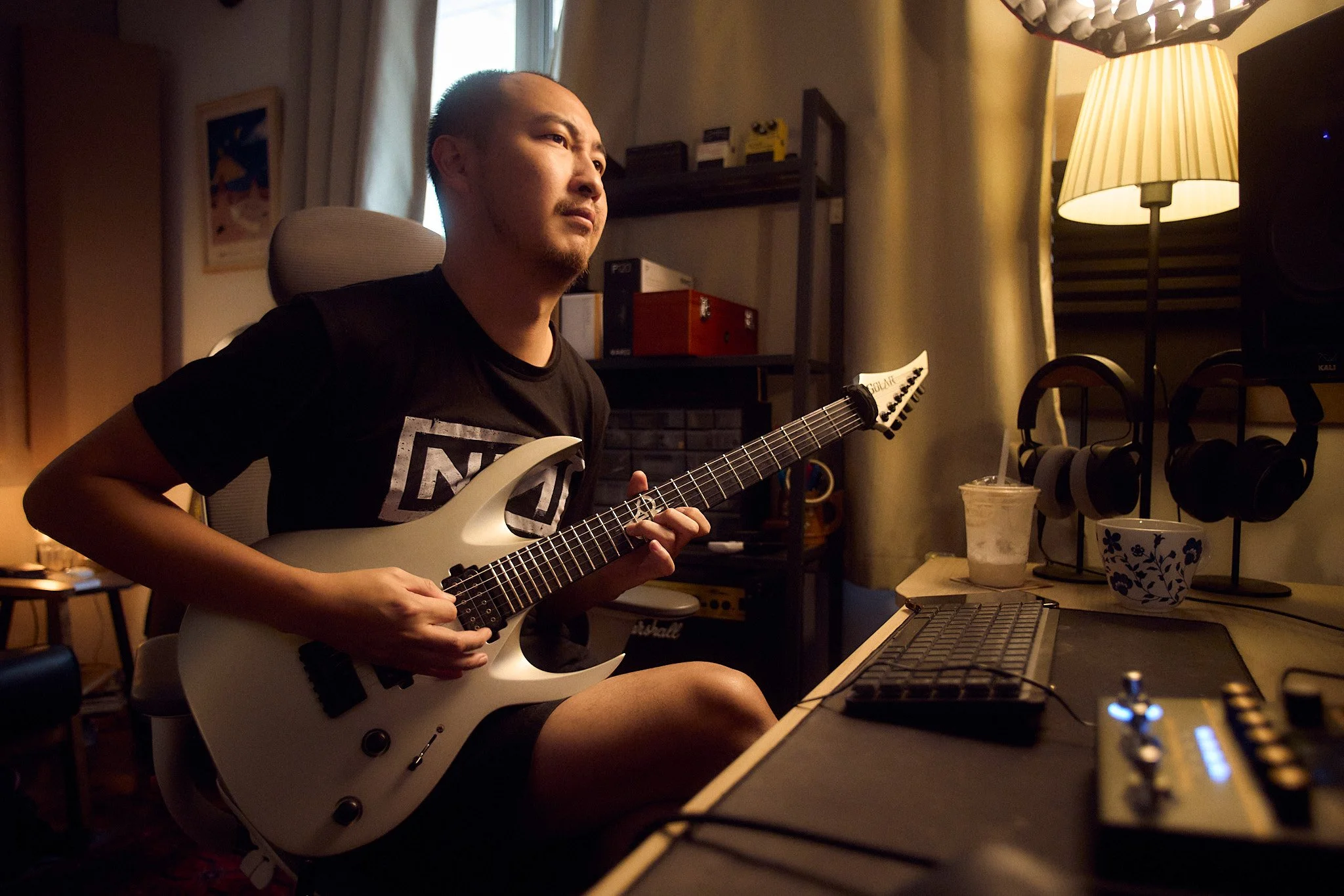 A man sitting in a music studio playing an electric guitar, with studio equipment and a lamp on a desk in the background.