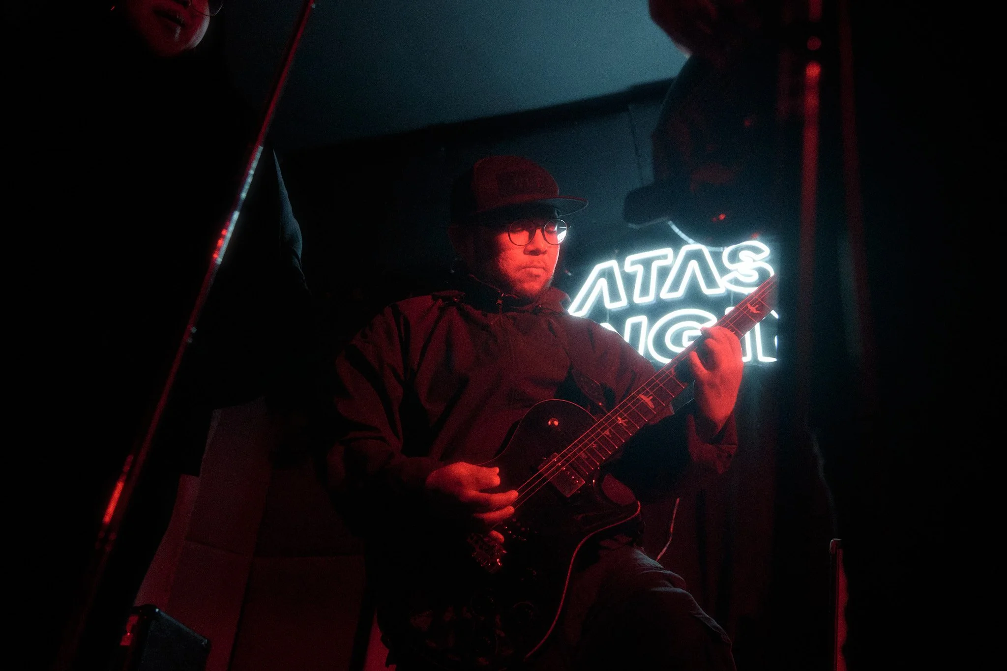 A man playing an electric guitar on stage, with a neon sign behind him that reads 'Atlas' and part of another word, in a dark setting.