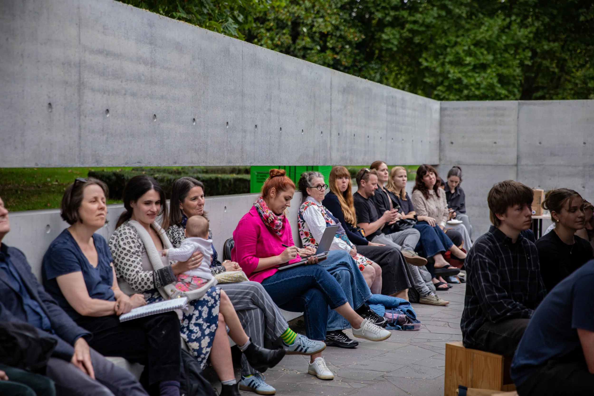 A group of people sitting on a bench, attending a participatory event. Some are using notebooks or laptops, and one person is holding a small child.