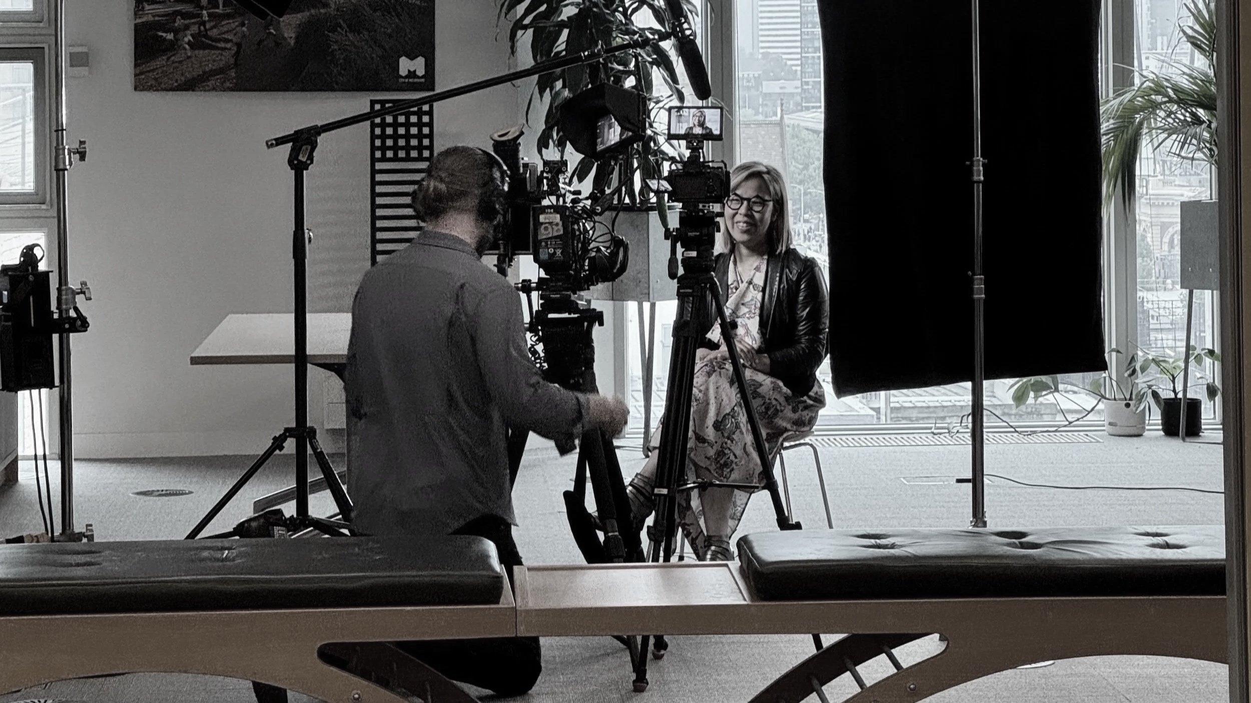 A woman, urbanist Jocelyn Chiew, being recorded in a studio with cameras, lights, and curtains. She is smiling, wearing glasses, a patterned dress, and a leather jacket, seated in front of a large window with plants outside.