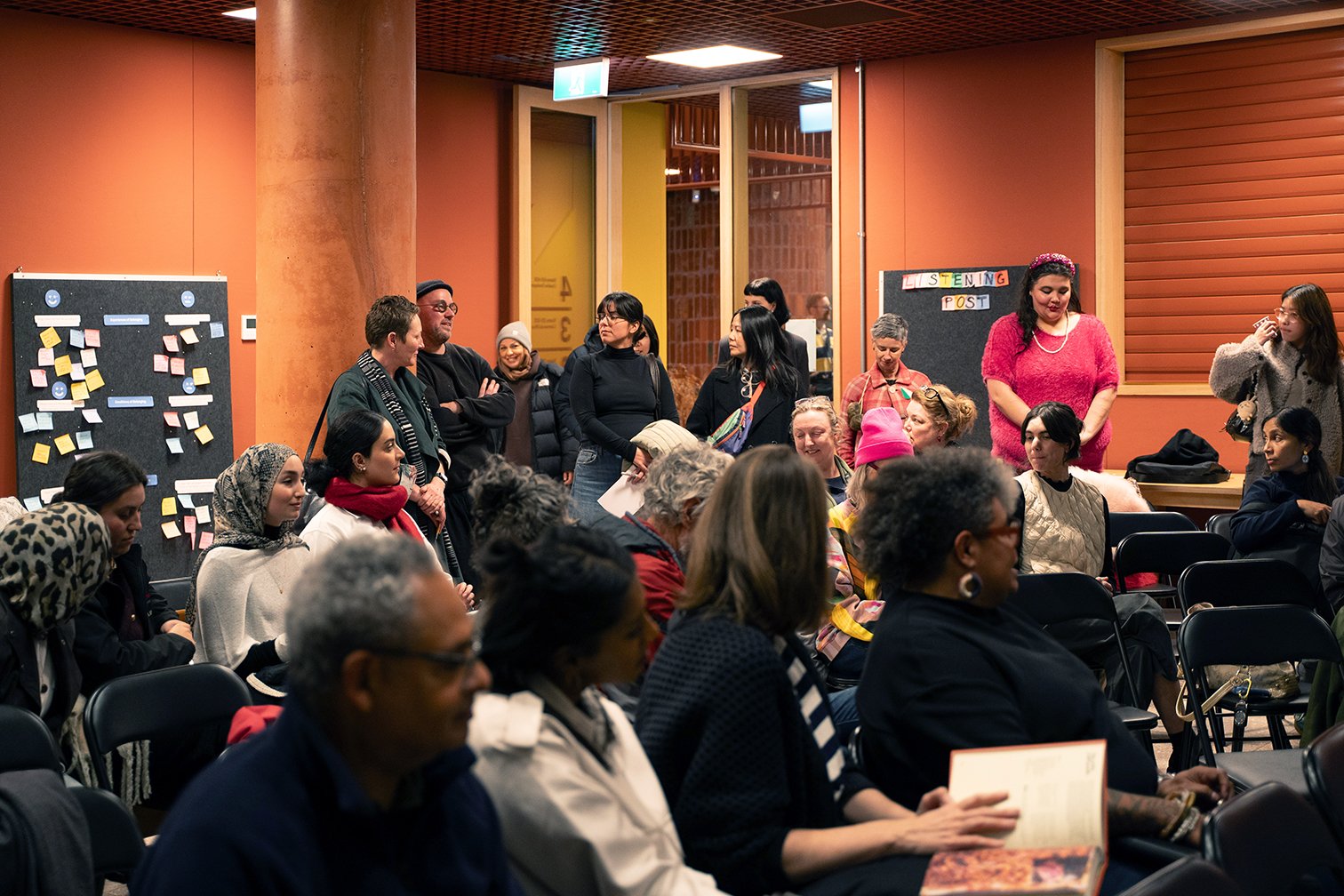 People gathered for a workshop on The Atlas of Belonging. Some people are seated, while others stand, listening or engaging. There are self-supporting felt boards in the background with handwritten notes contributed by participants.