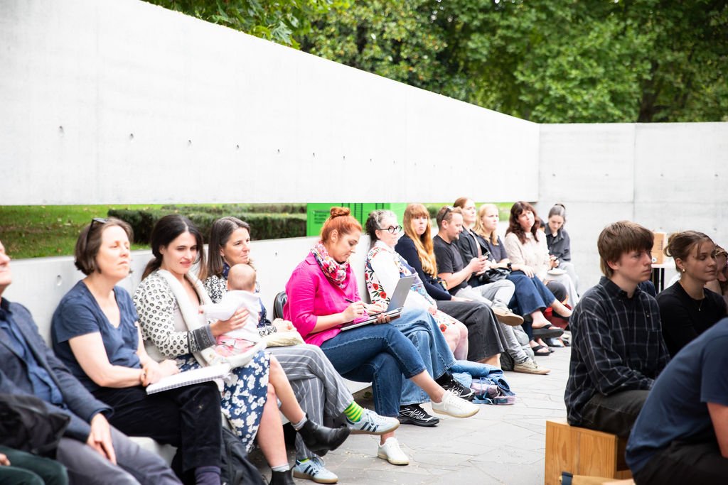 A group of people sitting outdoors on a concrete curved bench, at Tadao Ando's MPavilion, participating in a panel discussion and workshop as part of Shaping the Atlas of Belonging at MPavilion.