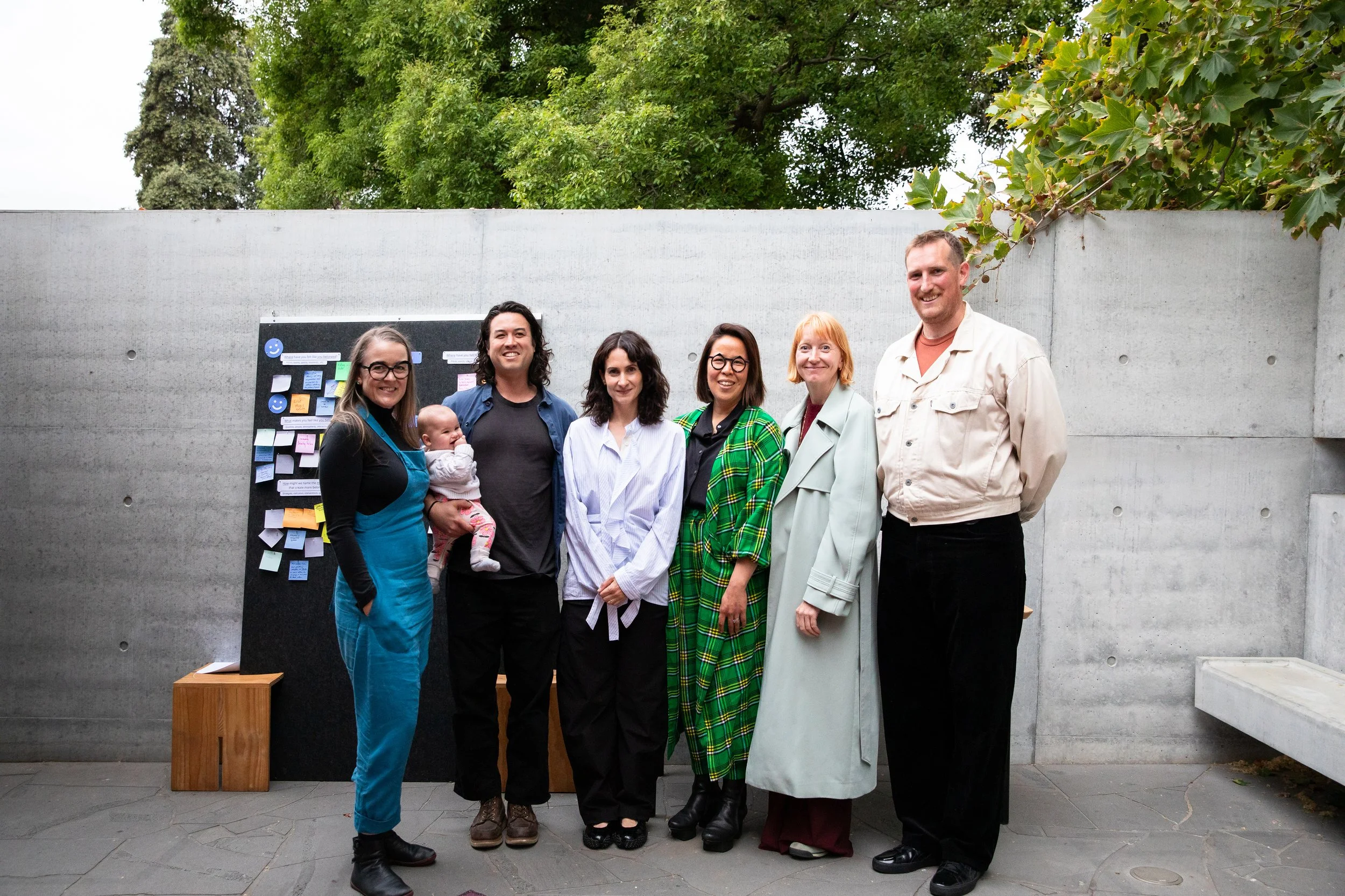 A group of seven people and a baby posing together outdoors in front of a concrete wall at MPavilion, with a display board to the left, some trees overhead, and a small bench at the left edge as part of Shaping the Atlas of Belonging at MPavilion.