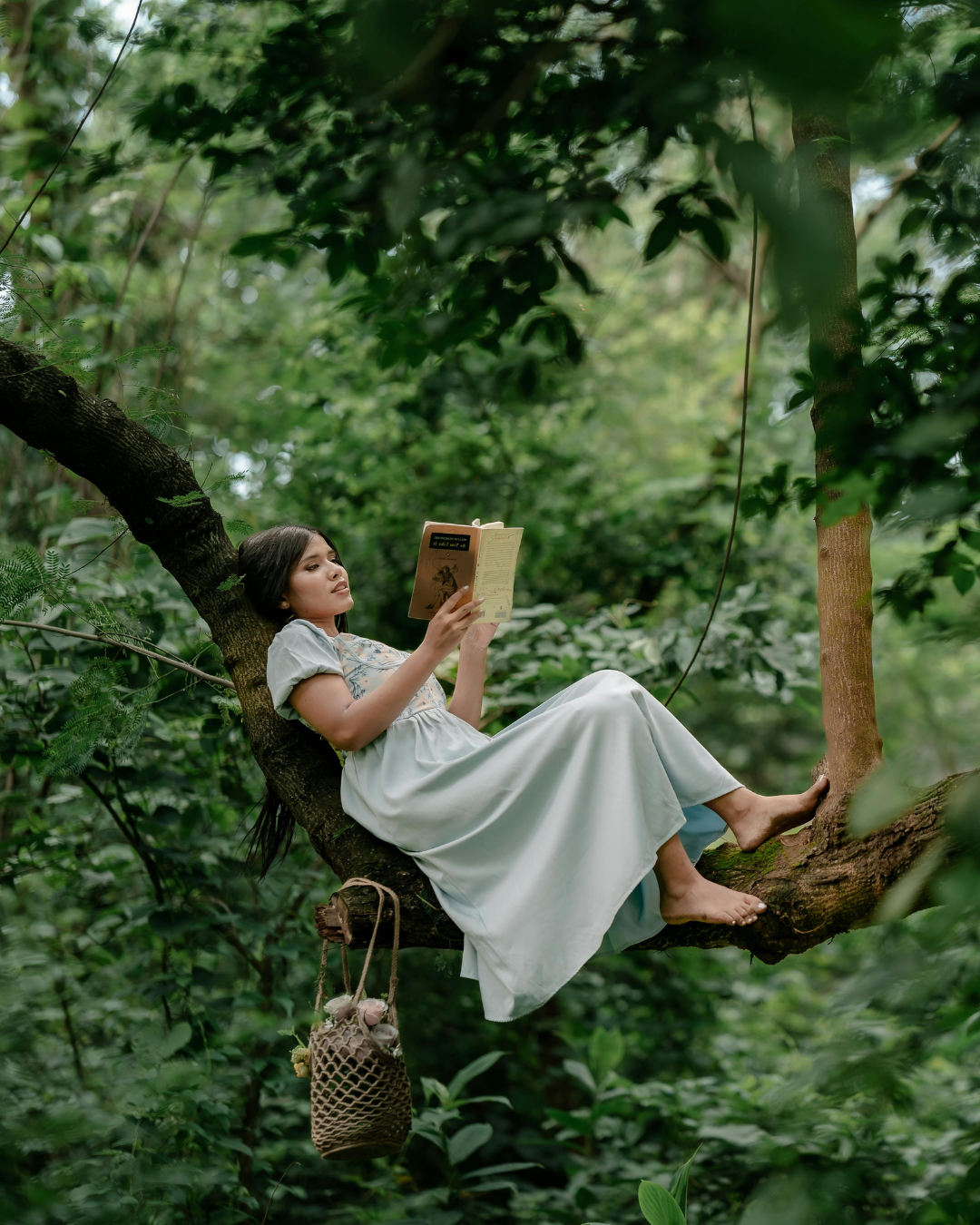 A woman in a white dress reclining on a tree branch in a lush, green forest, reading a book with a netted bag hanging from the branch.