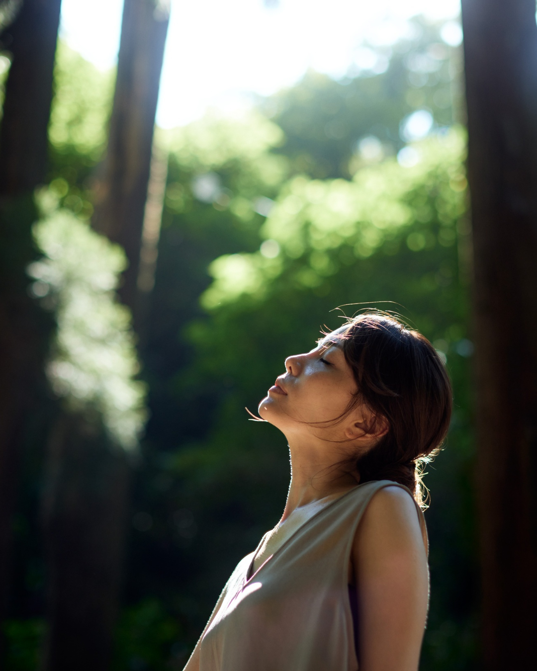 A woman with short dark hair standing in a forest with sunlight shining through the trees, her eyes closed and face turned slightly upward.