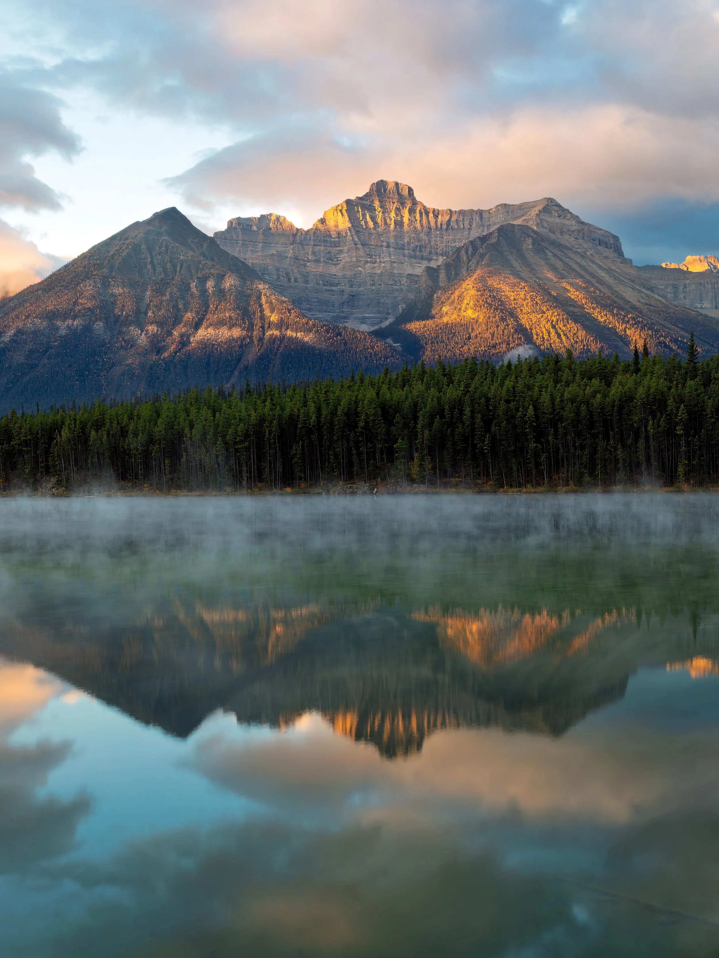 Sunlit mountain peaks reflected in a calm lake with a forest in the foreground and clouds in the sky.