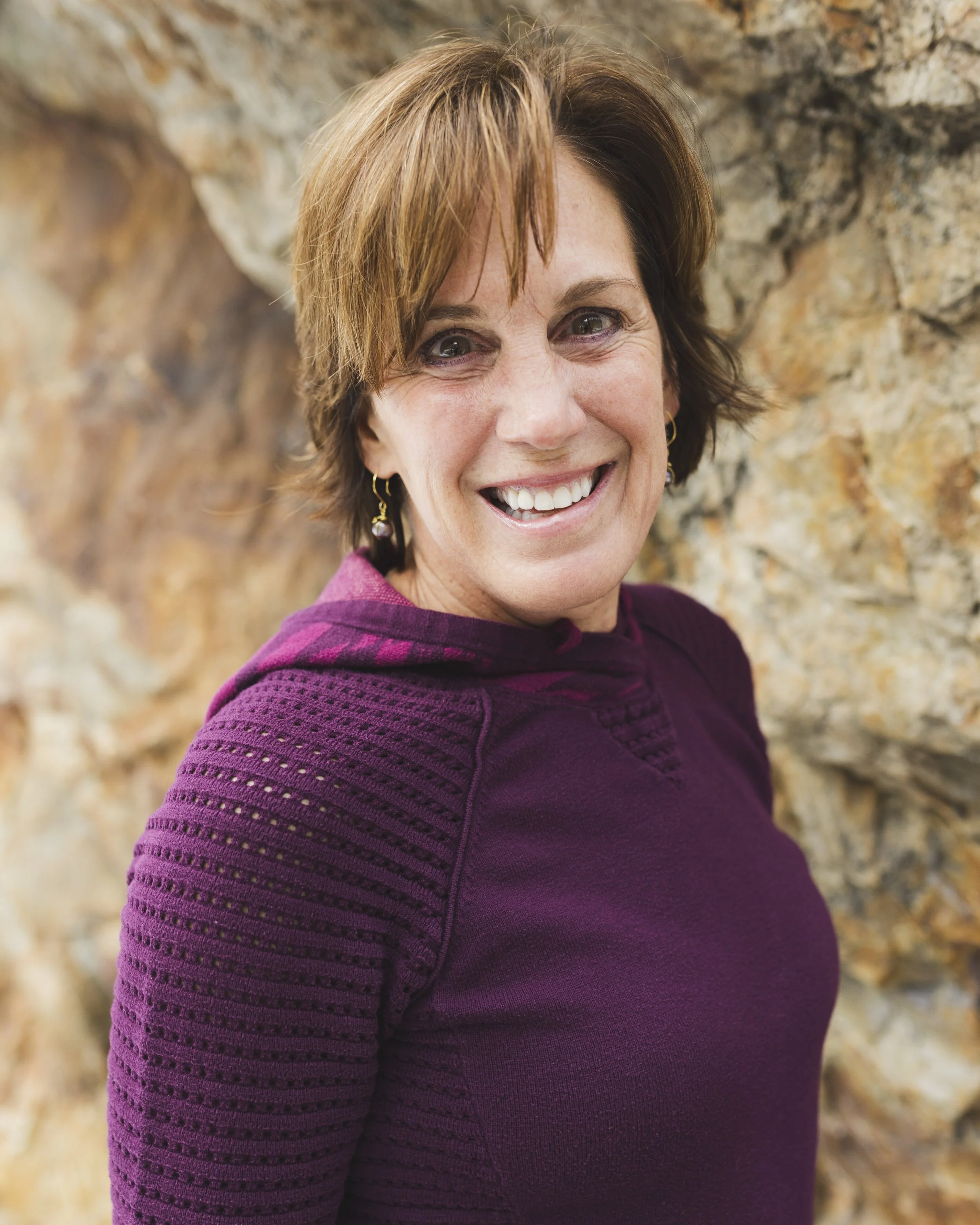 Close-up of a smiling woman with short brown hair, wearing a purple sweater, standing in front of a rocky background.
