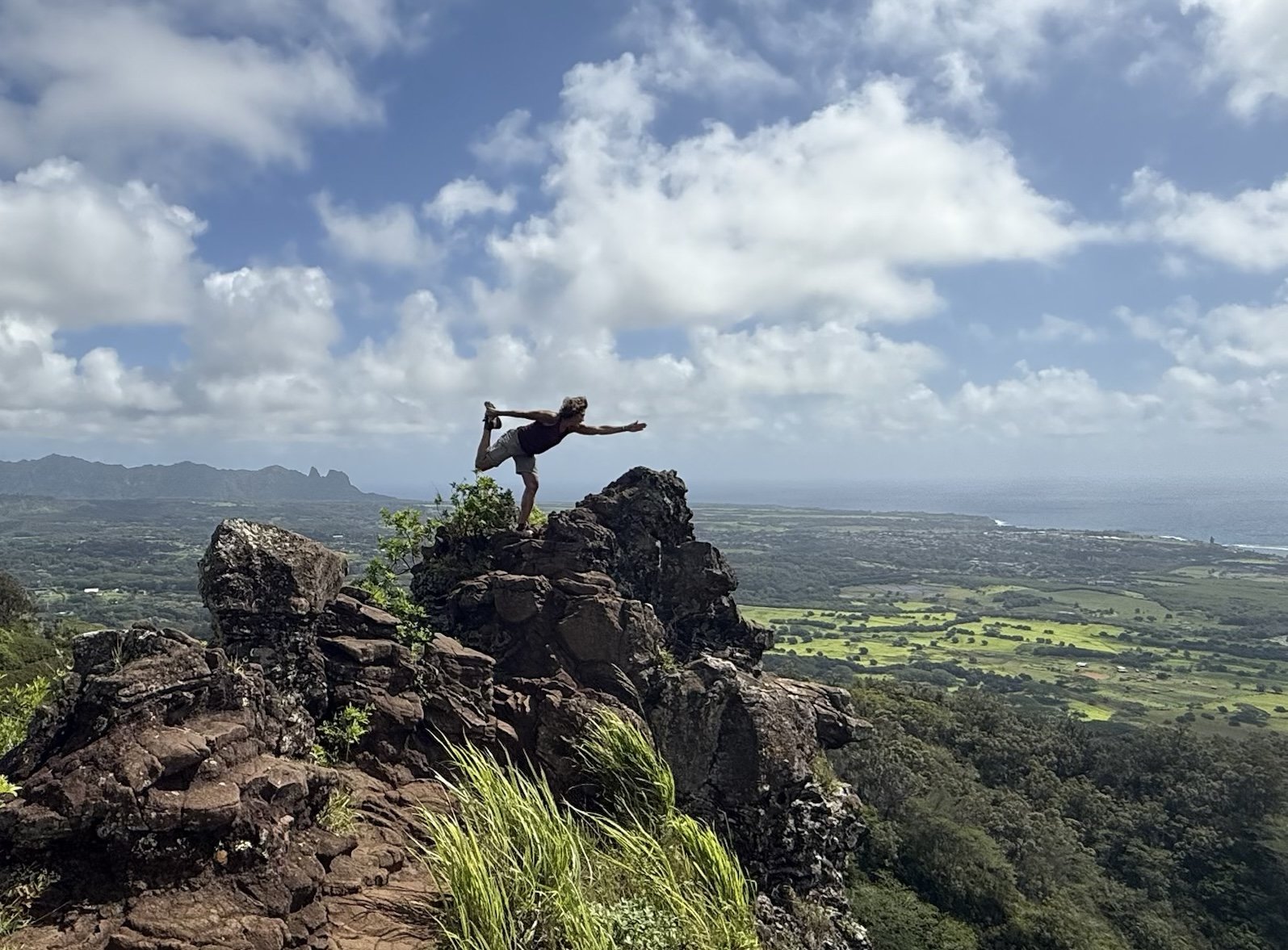 A person practicing yoga on a rocky mountain peak with a cityscape, ocean, and mountains in the background under a partly cloudy sky.