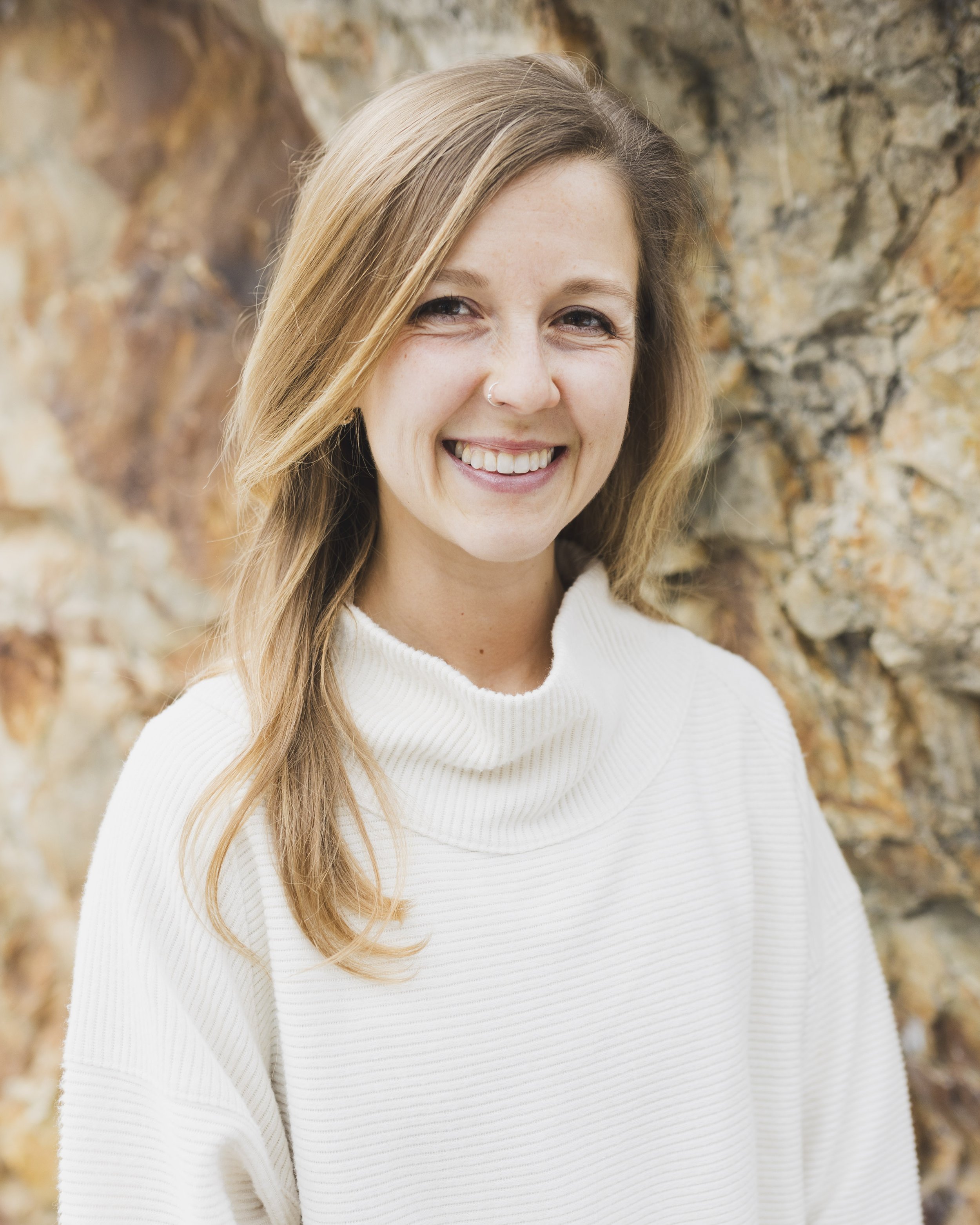 A smiling woman with long light brown hair wearing a white turtleneck sweater, standing in front of a rocky background.