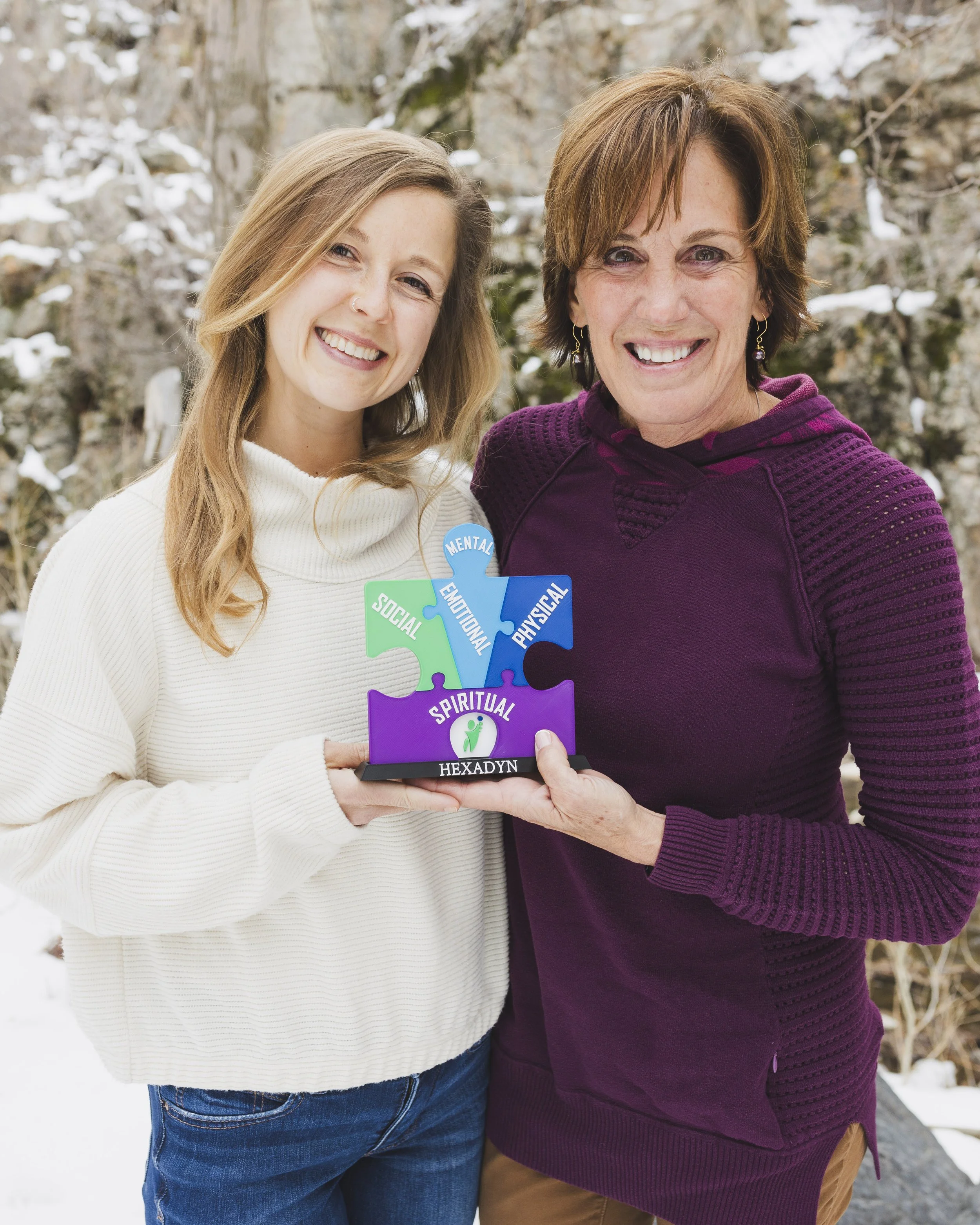 A young woman and an older woman outdoors in snowy terrain, smiling and holding a colorful award with puzzle pieces labeled 'Social,' 'Mental,' 'Emotional,' 'Physical,' and 'Spiritual,' and the name 'HEXADYN' at the bottom.