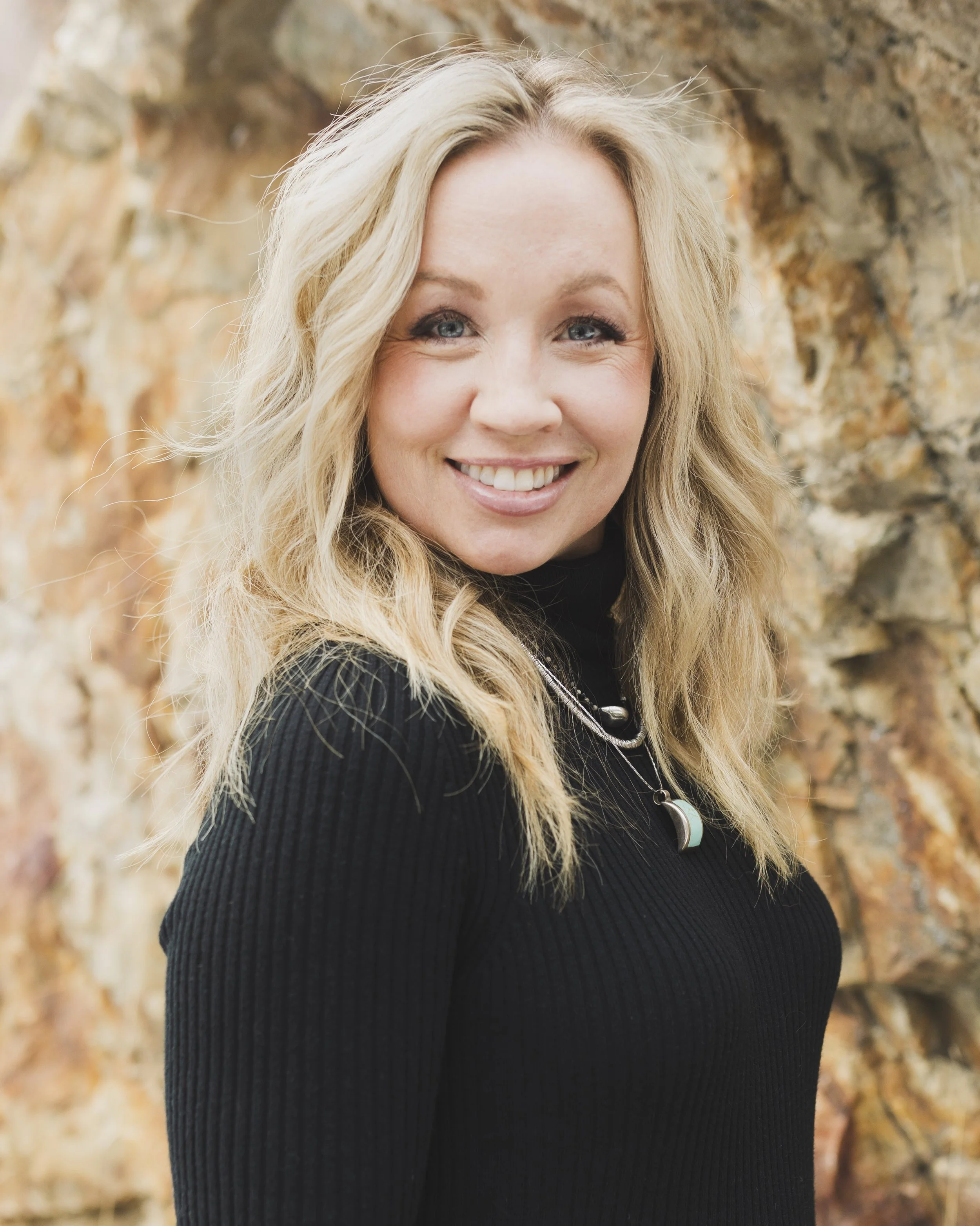 A smiling woman with long, wavy blonde hair posing outdoors with a stone wall in the background.