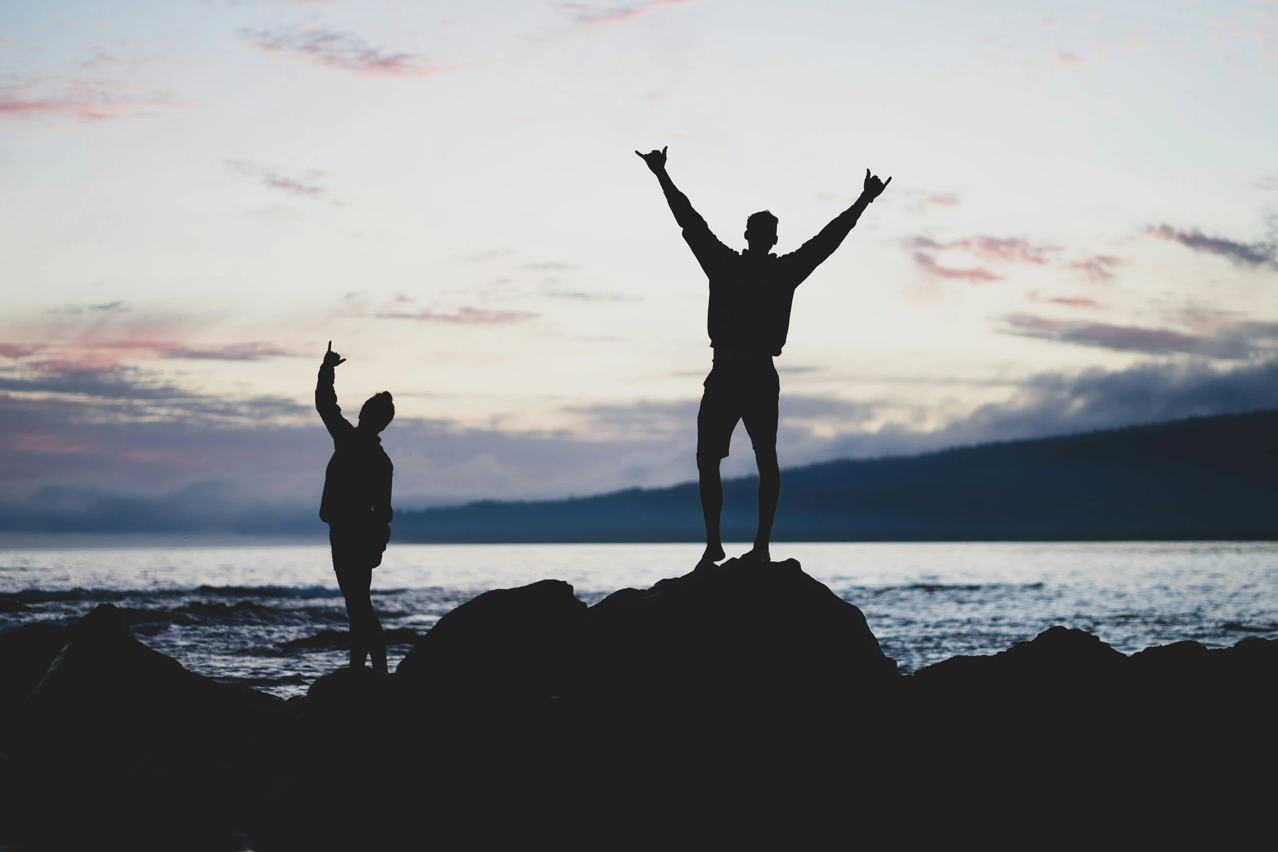 Silhouettes of two people standing on rocks near water at sunset, one with arms raised and the other with one arm raised.
